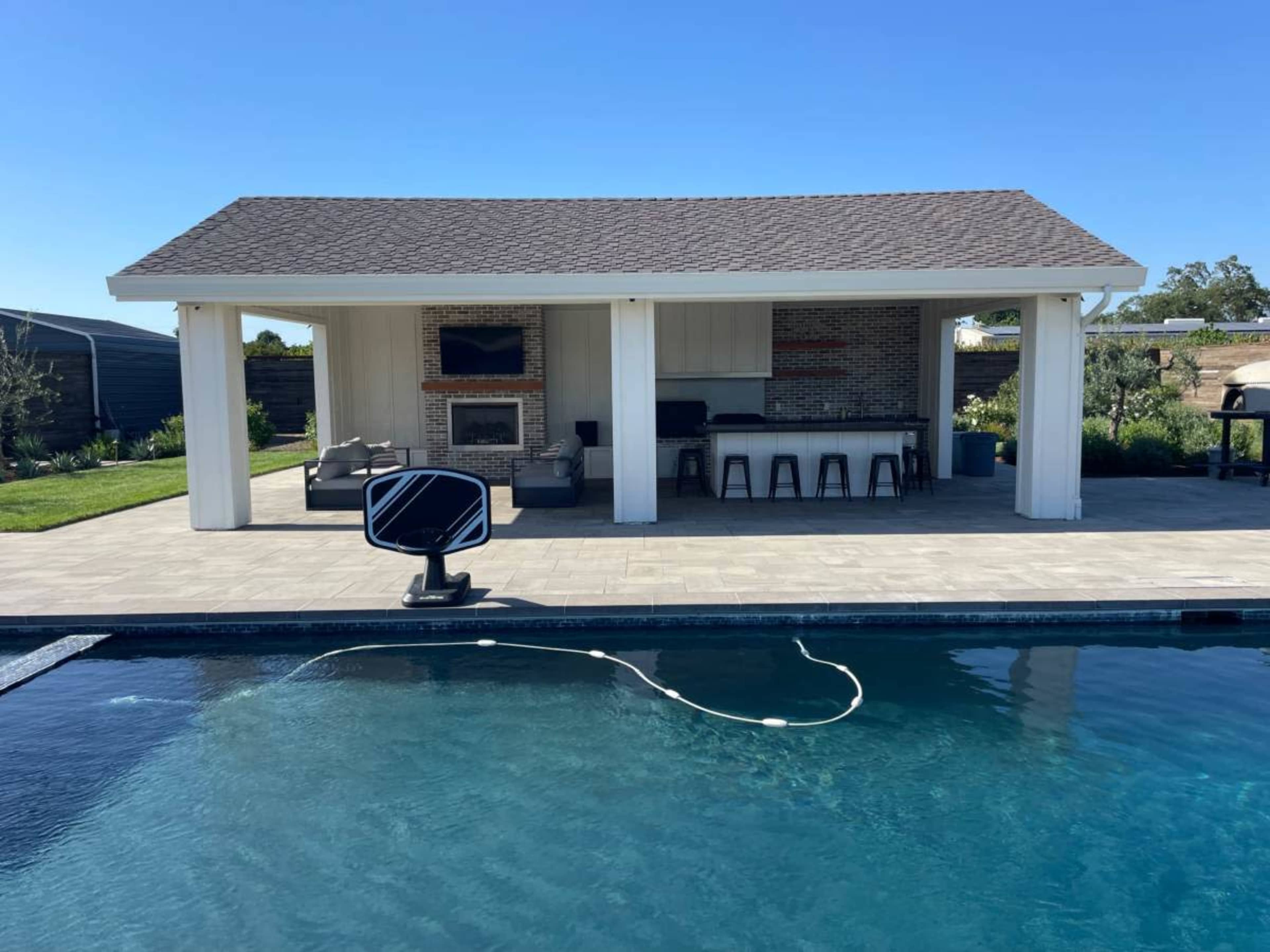 The image shows a modern outdoor pool area with a covered patio that features a fireplace, seating area, and an outdoor kitchen.