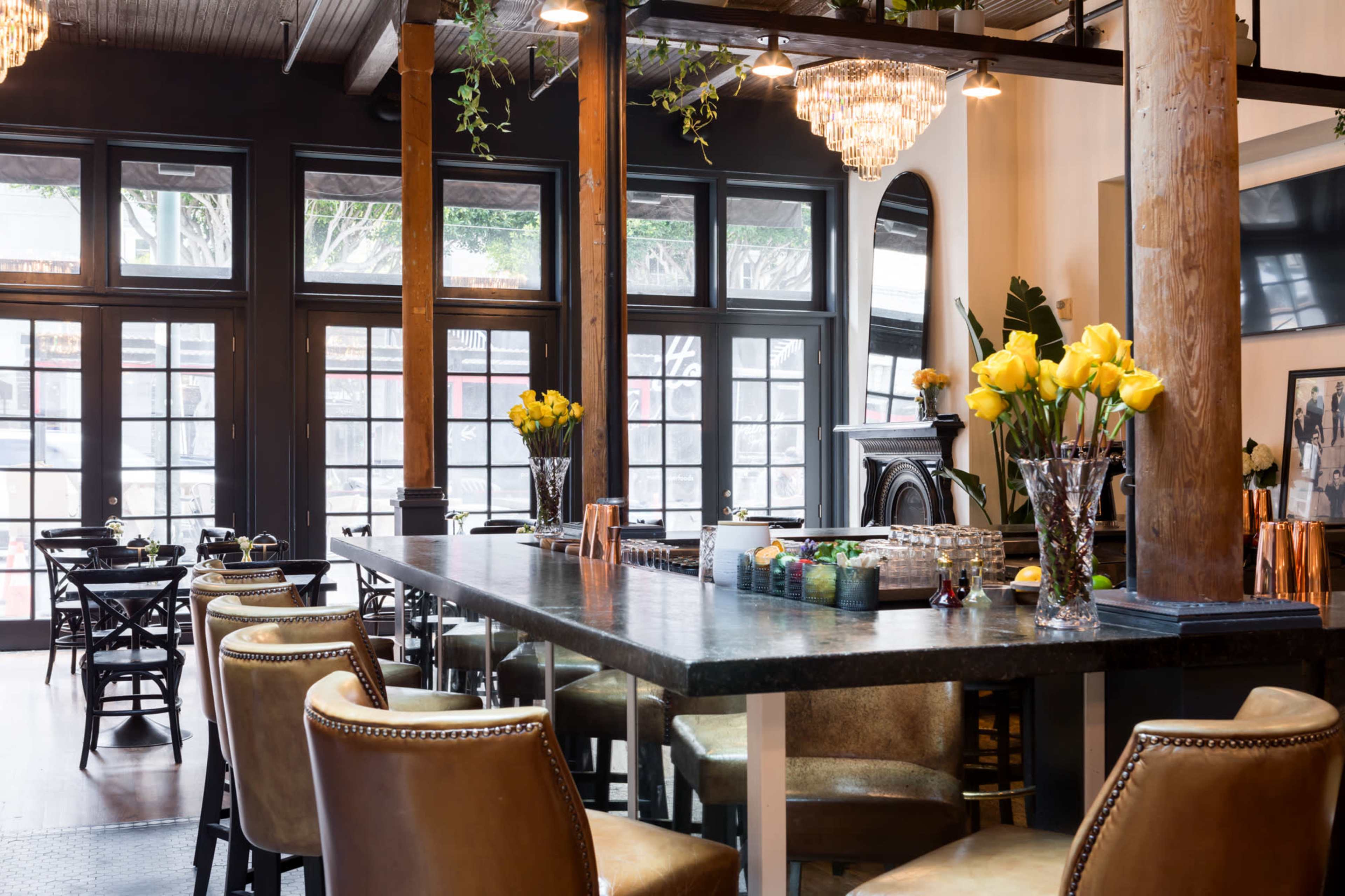 A stylish interior of a bar or restaurant features a long table with flowers, surrounded by chairs and large windows letting in natural light.