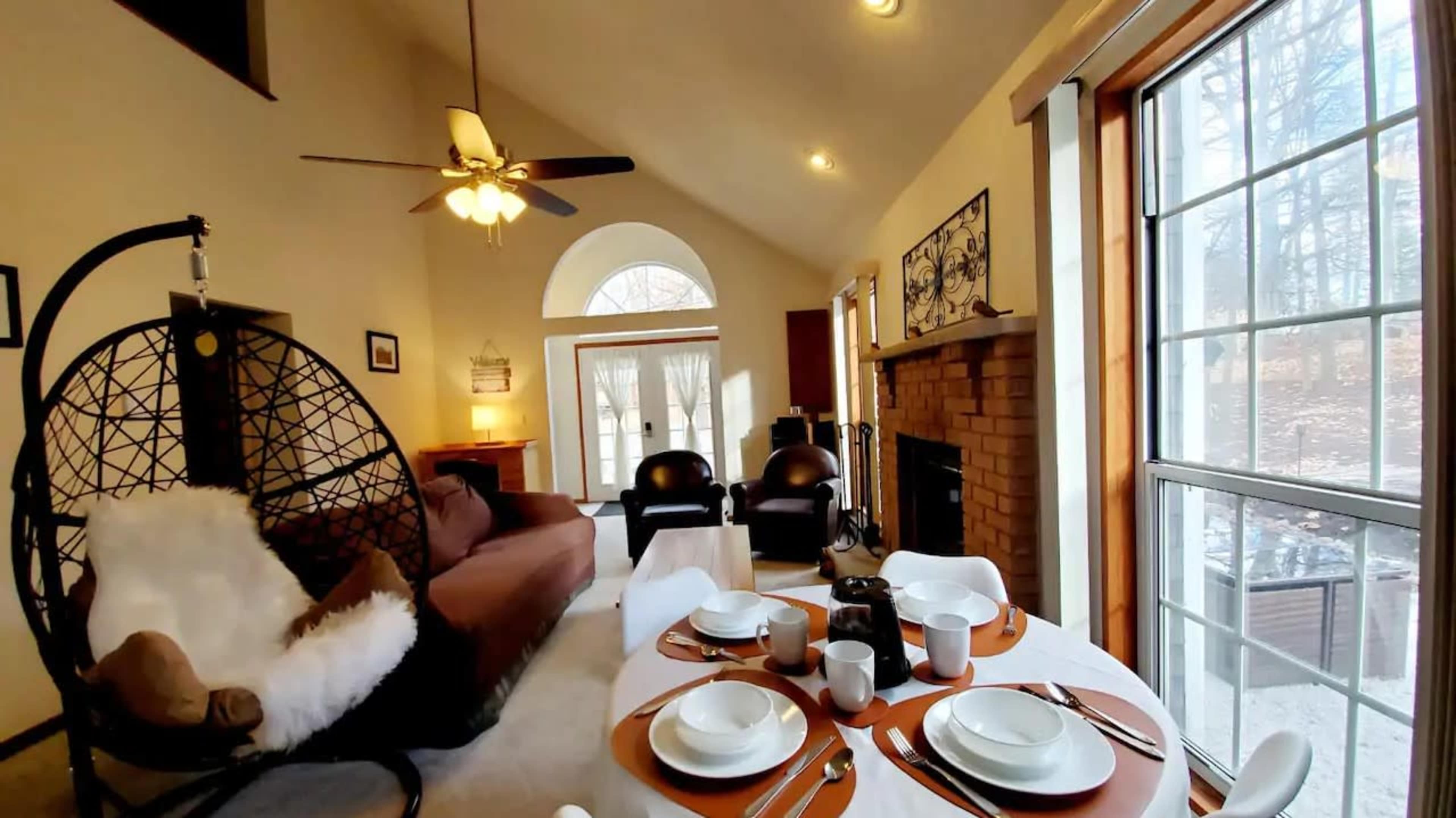 The image shows a cozy living room with a hanging chair, a coffee table set for a meal, and large windows allowing natural light to enter.