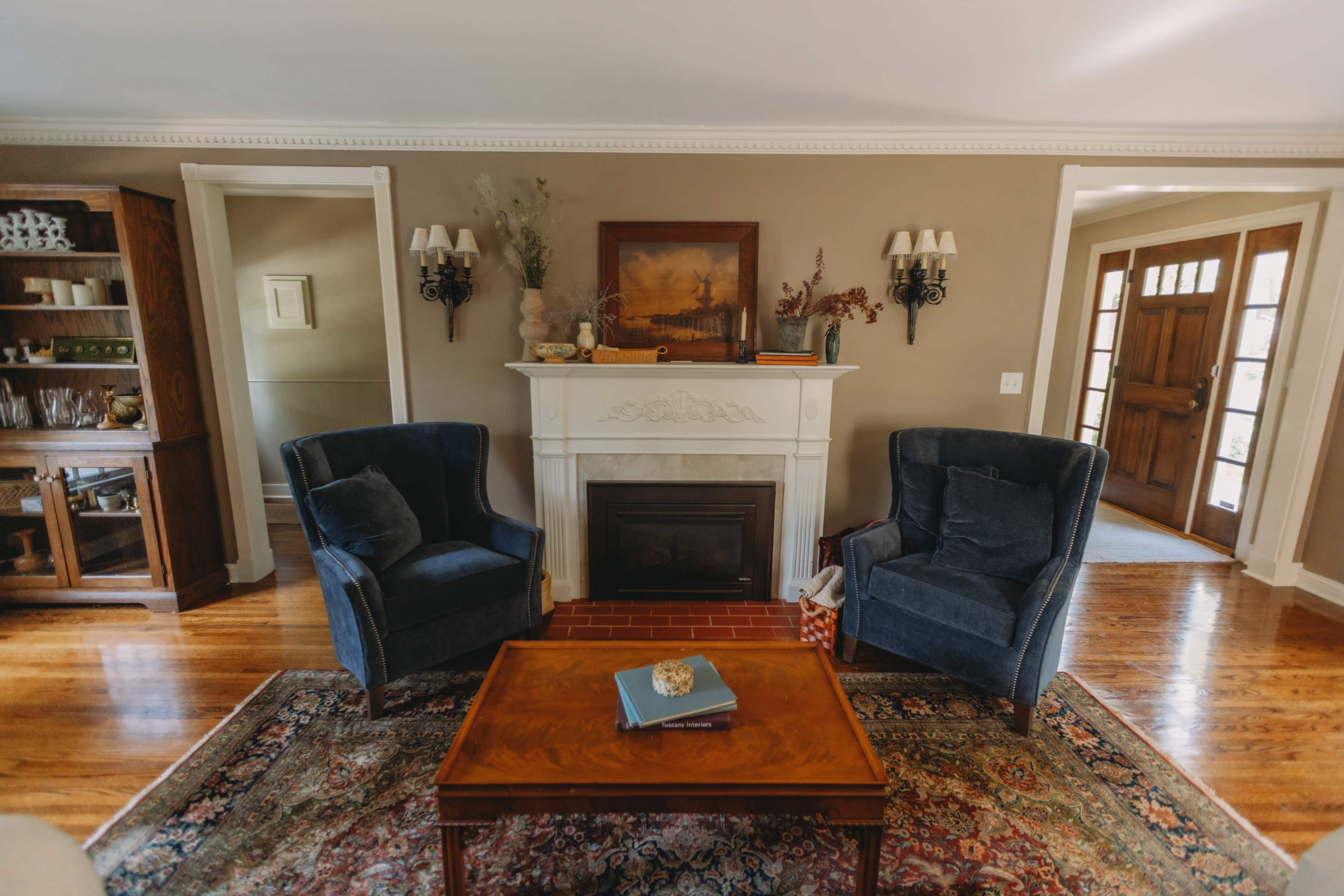 The image shows a cozy living room with two blue armchairs positioned near a fireplace and a wooden coffee table on a patterned area rug.