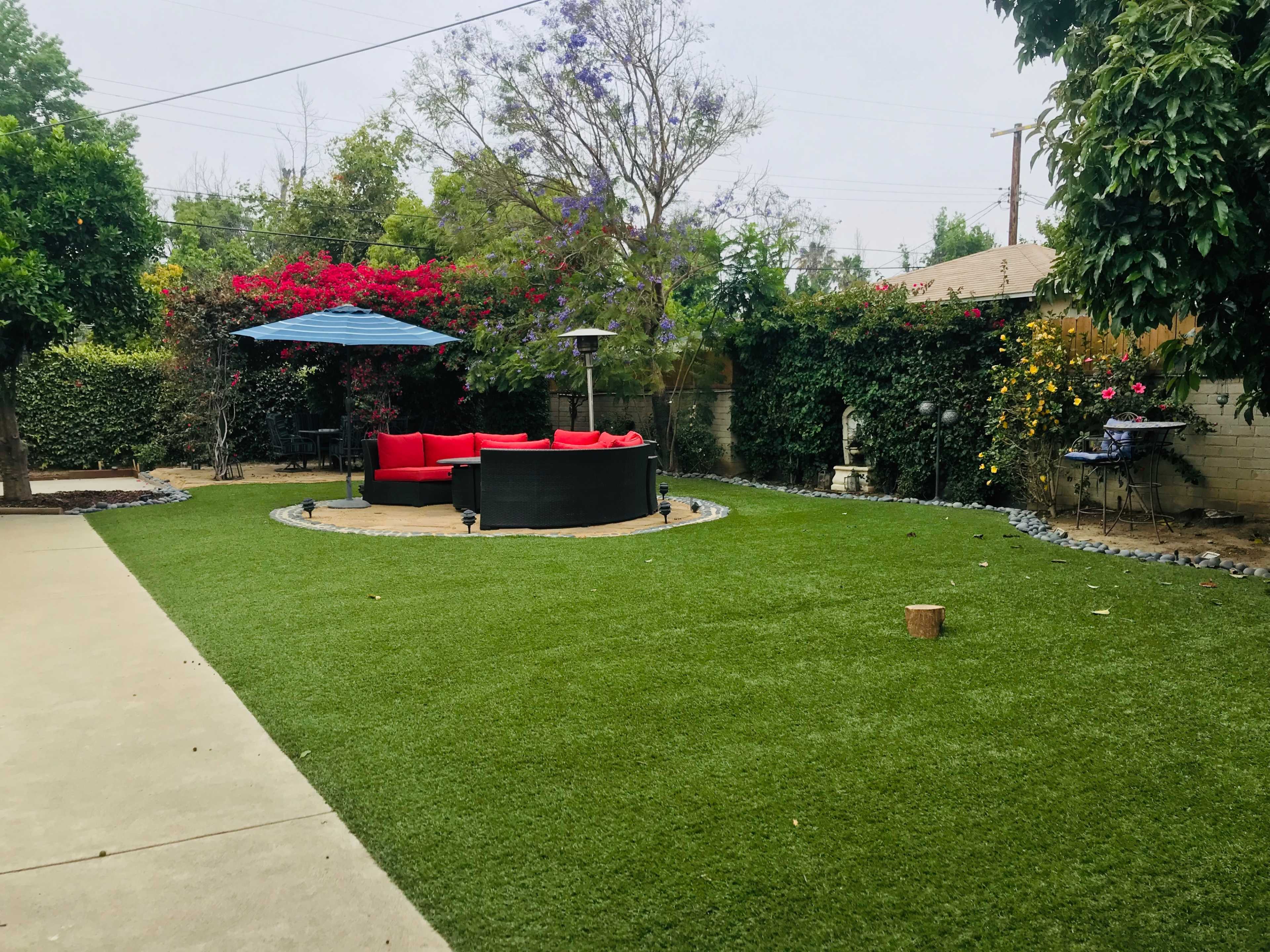 A landscaped backyard features a circular area of artificial turf with a black seating arrangement and red cushions, surrounded by greenery and flowering plants.