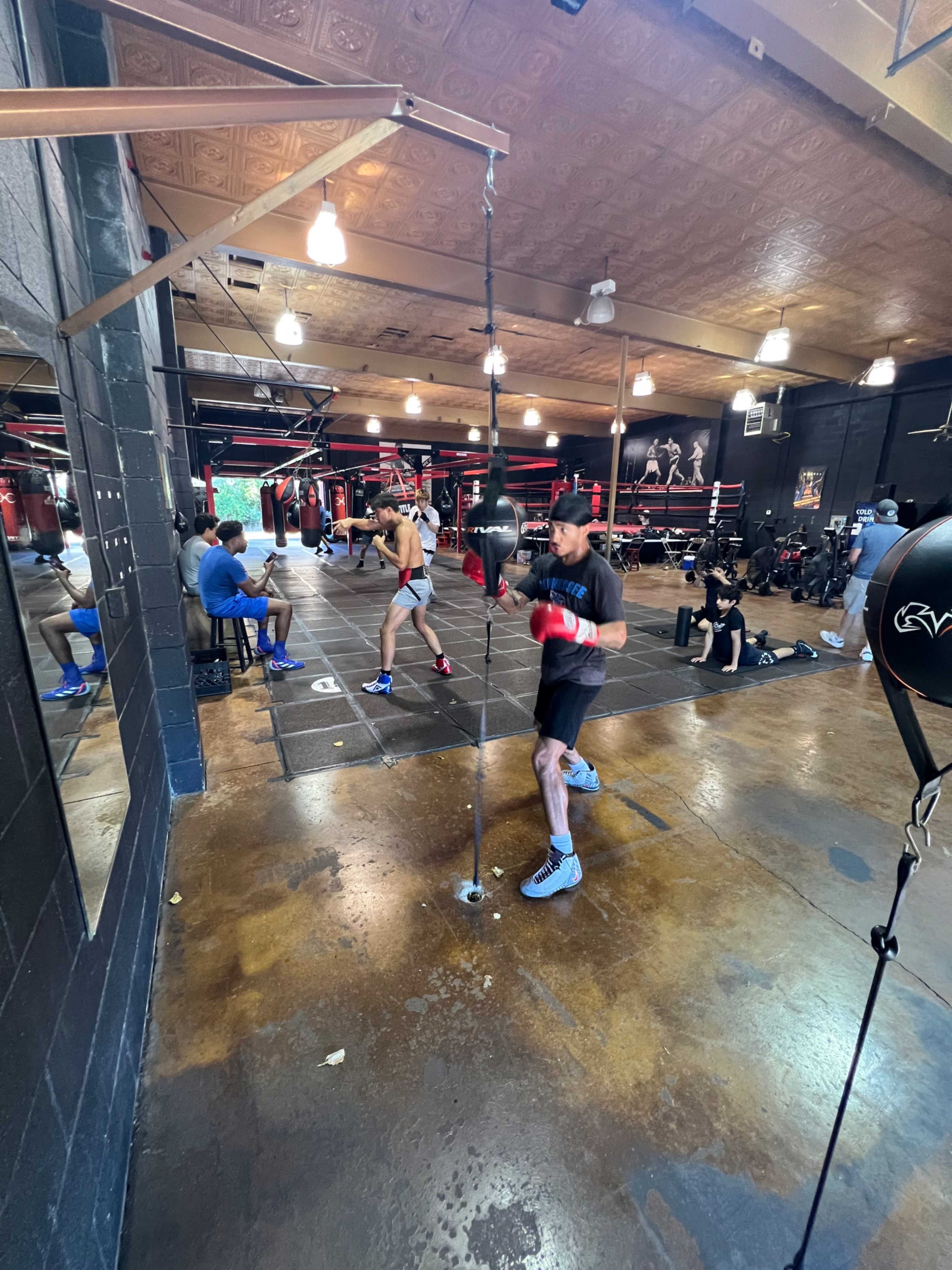 A boxer practices on a speed bag in a gym while others train or watch nearby.