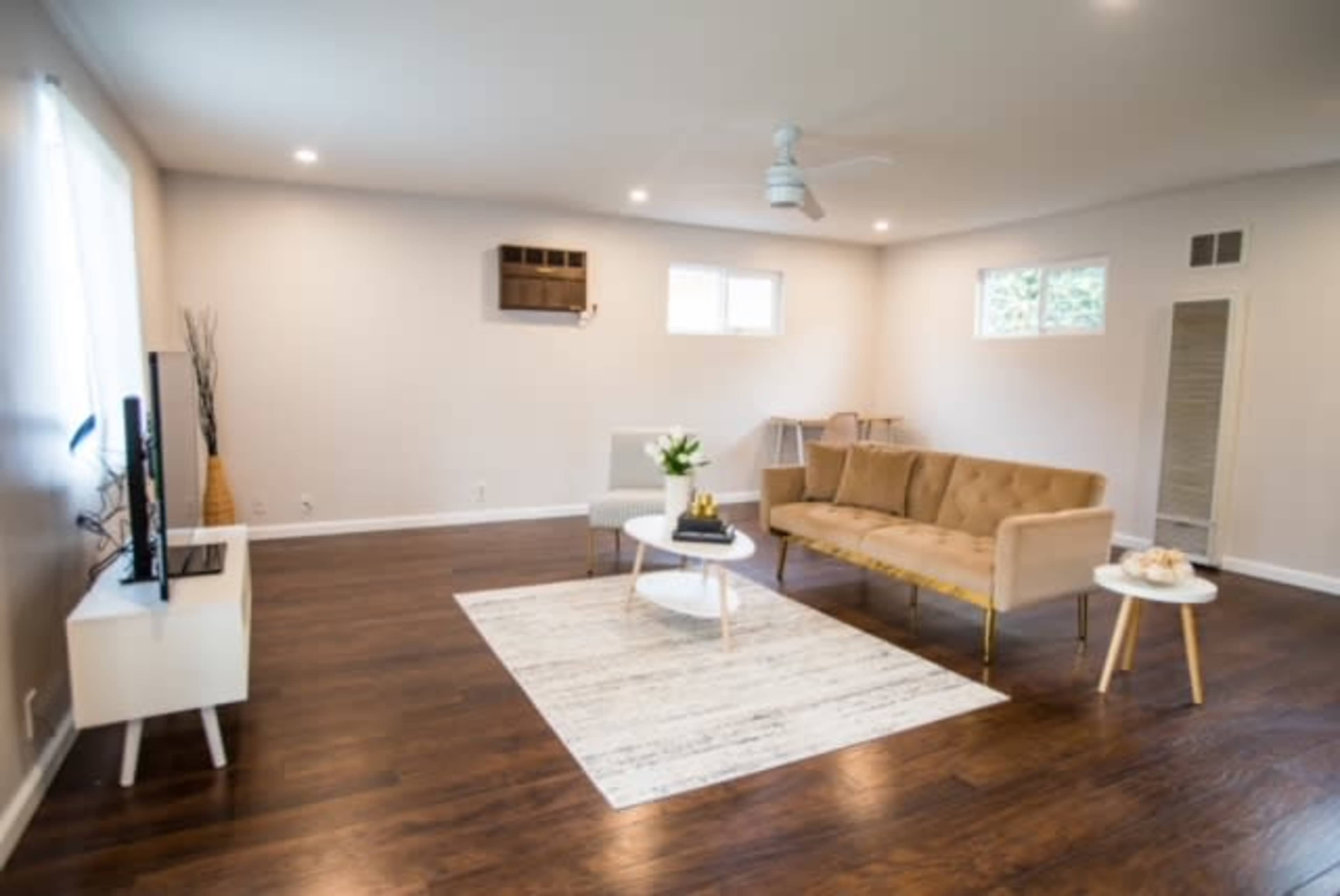 A neatly arranged living room features a beige sofa, a round coffee table, a television stand, and a light rug on dark wooden flooring.
