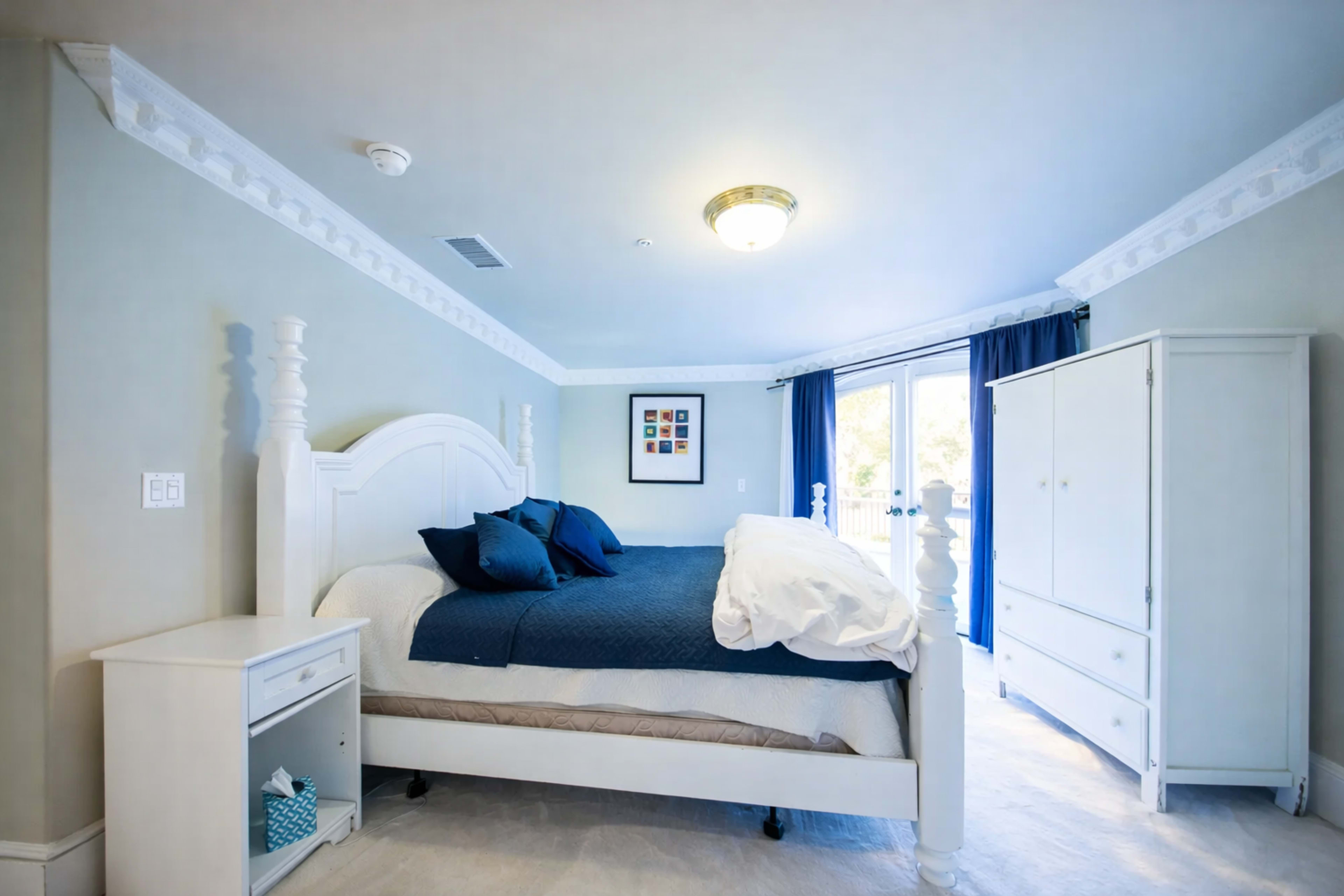 A bedroom featuring a white bed with blue bedding, a white nightstand, a dresser, and large windows with blue curtains.
