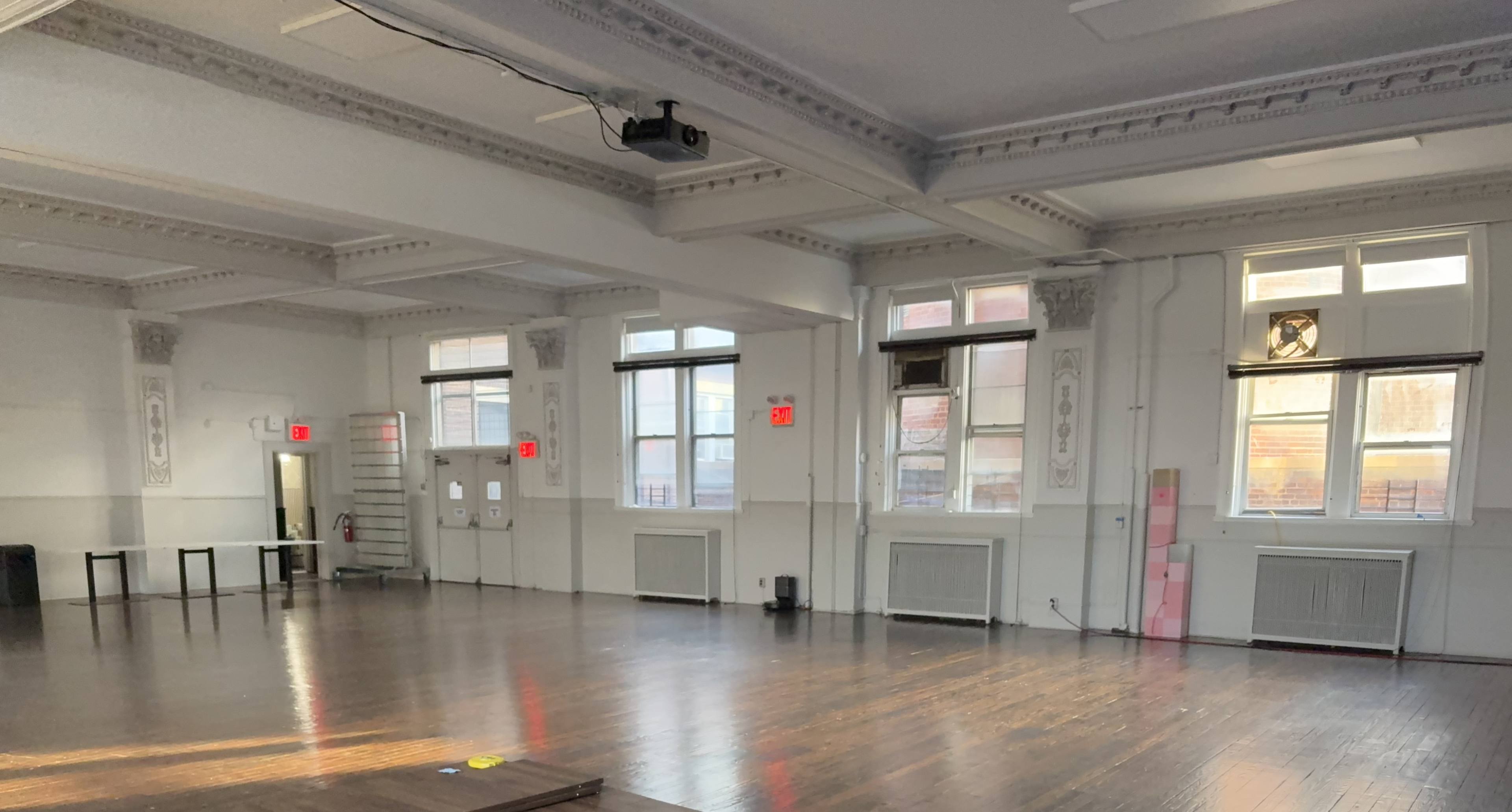 The image shows a spacious, well-lit room with wooden floors, large windows, and exposed pipes along the ceiling.