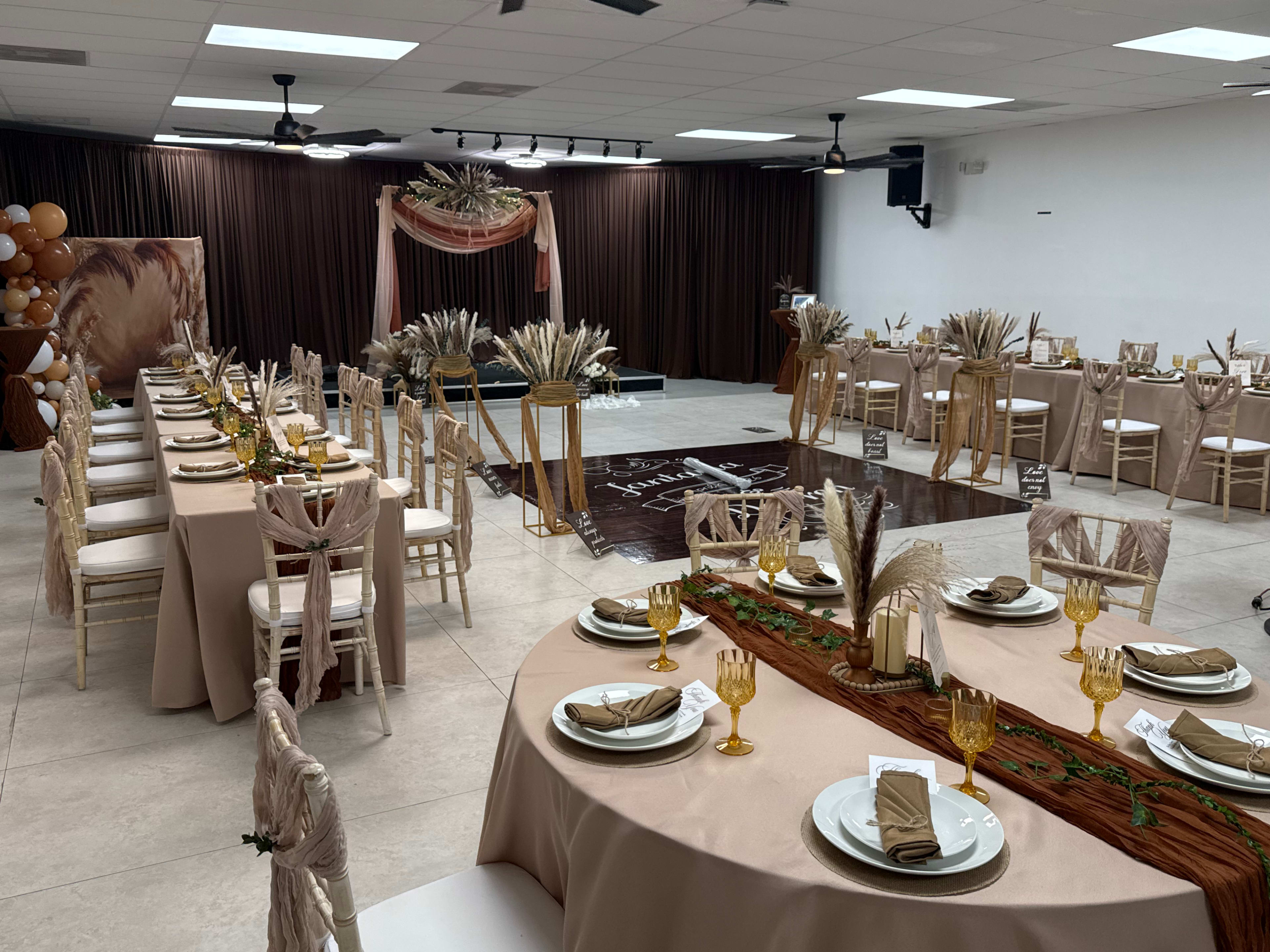 The image shows a decorated event space with elegantly set tables featuring neutral-colored tablecloths, centerpieces of dried flowers, and golden tableware, along with a backdrop of balloons and drapery.