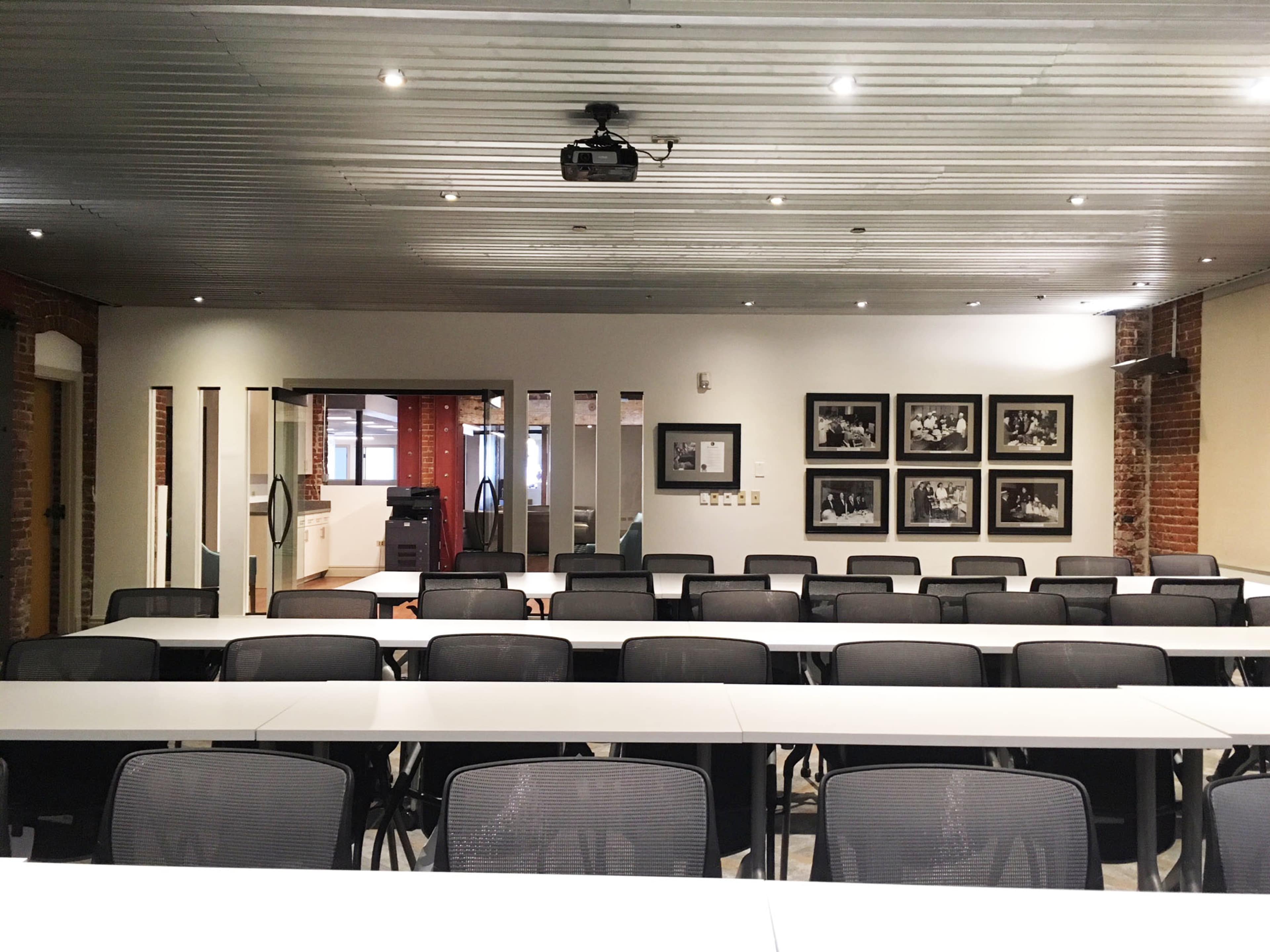 A modern conference room featuring rows of tables with black mesh chairs, a ceiling-mounted projector, and framed photographs on the walls.