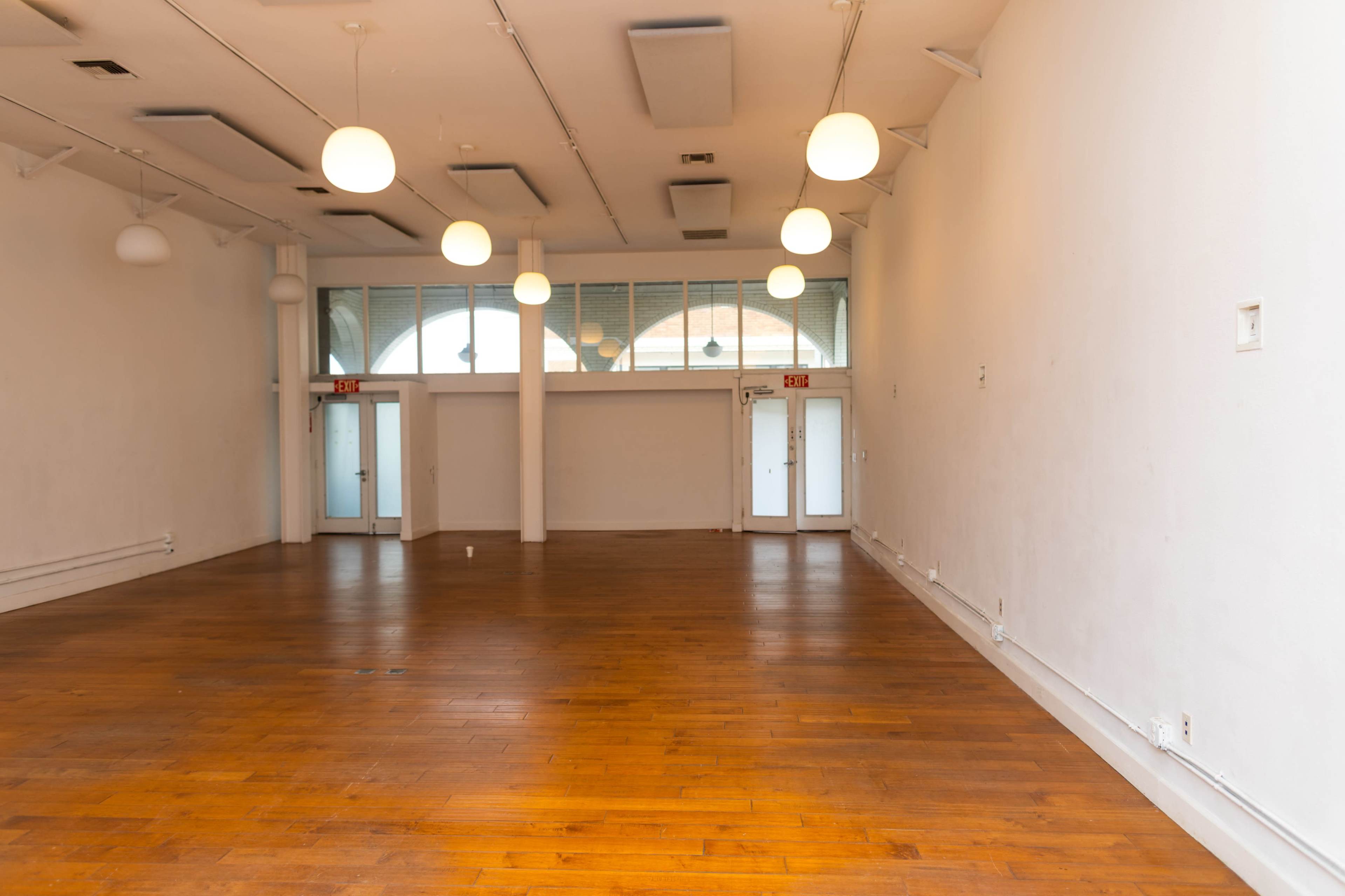 The image shows an empty indoor space with wooden flooring, large windows, and pendant lights hanging from the ceiling.