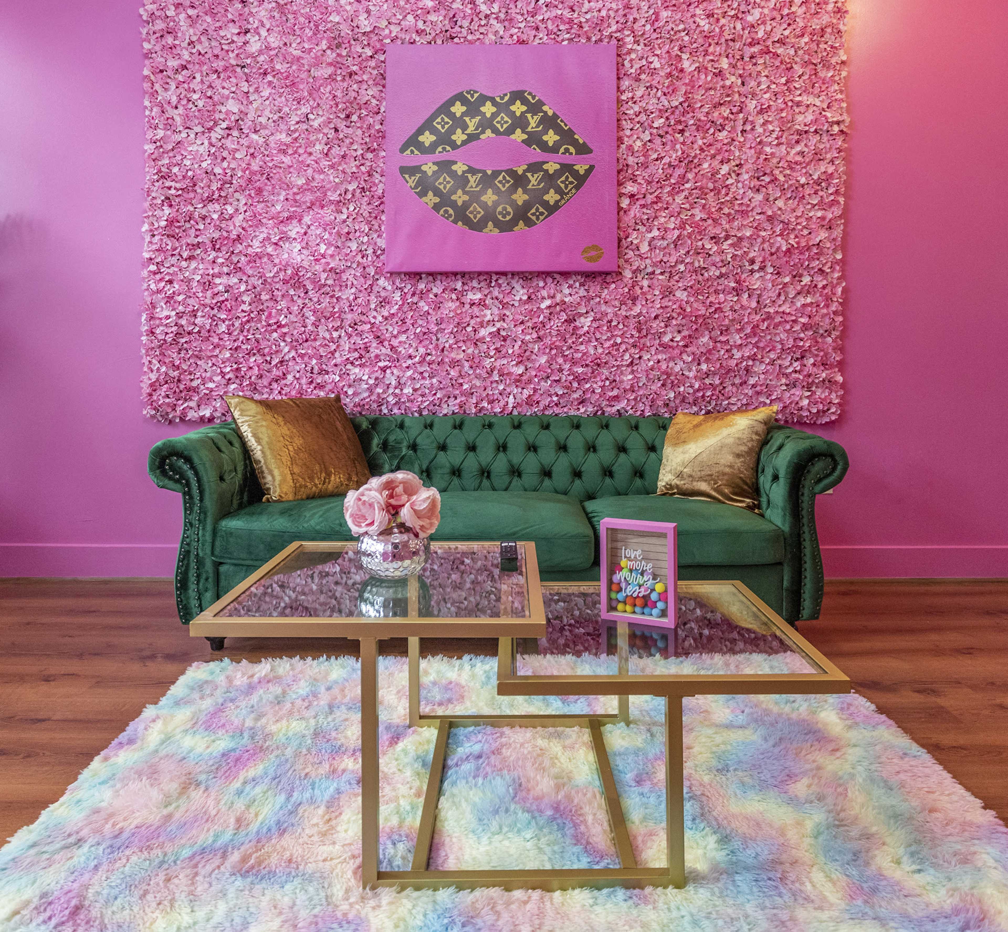 A green tufted sofa in front of a pink floral wall, with a glass coffee table and colorful decor items on a fluffy pastel rug.