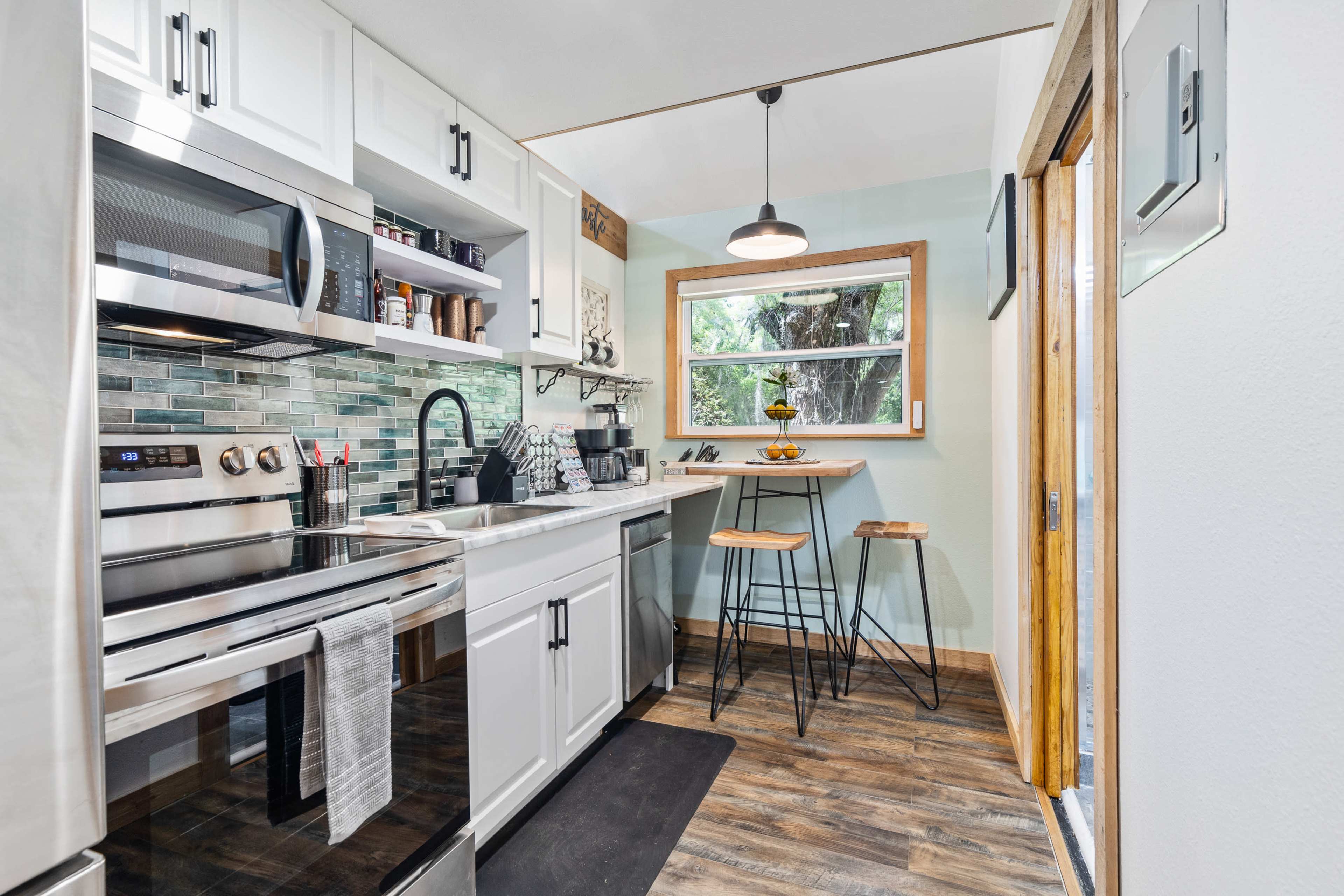 A compact kitchen featuring white cabinetry, stainless steel appliances, and a small dining area with two stools.