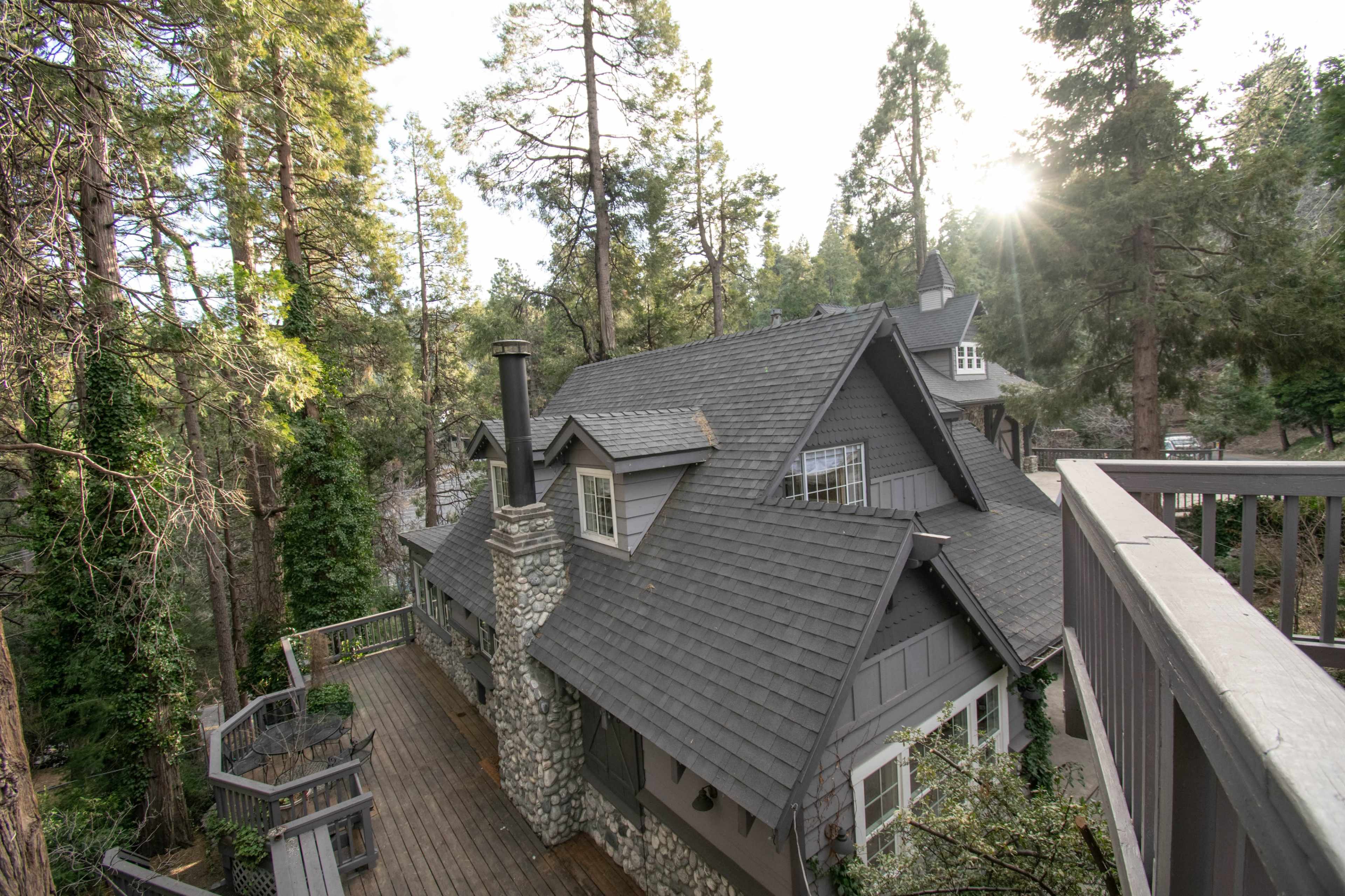 A gray cottage with a stone chimney is nestled among tall pine trees, featuring a wooden deck.