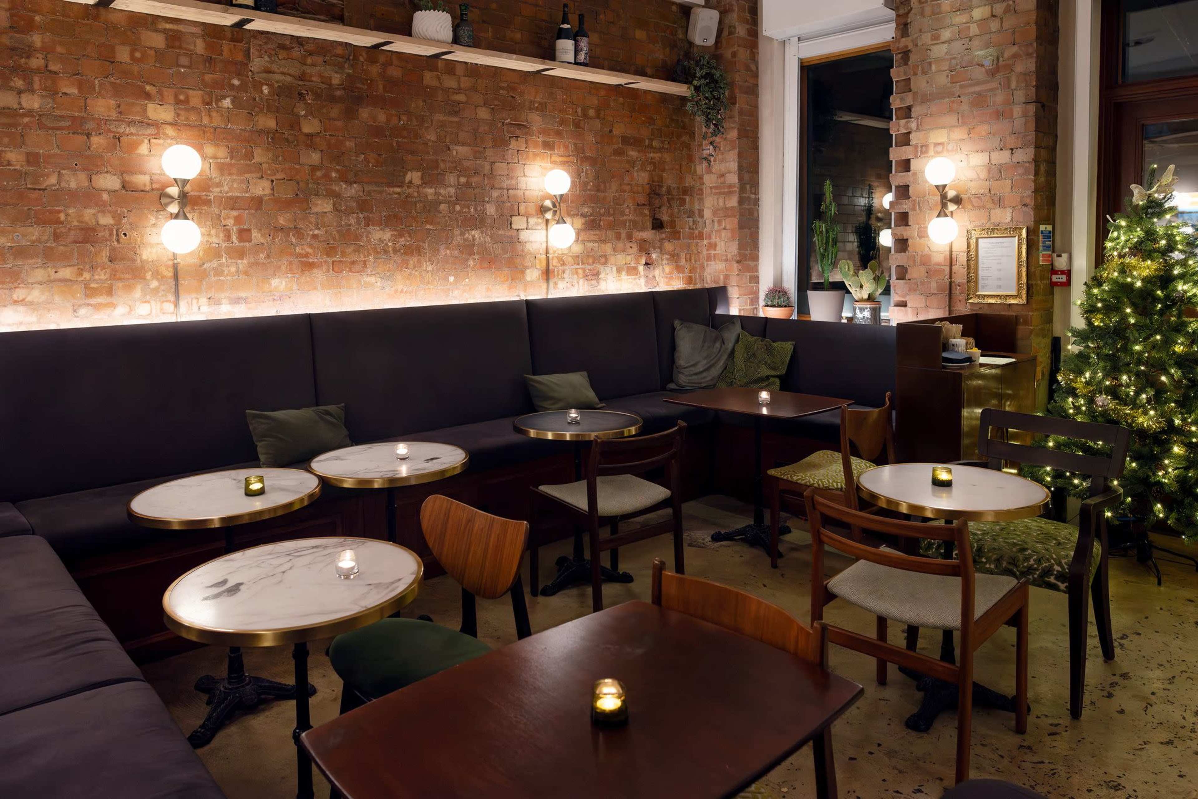 The image shows a cozy restaurant interior with exposed brick walls, dark seating, small tables, and decorative lights, along with a Christmas tree in the corner.