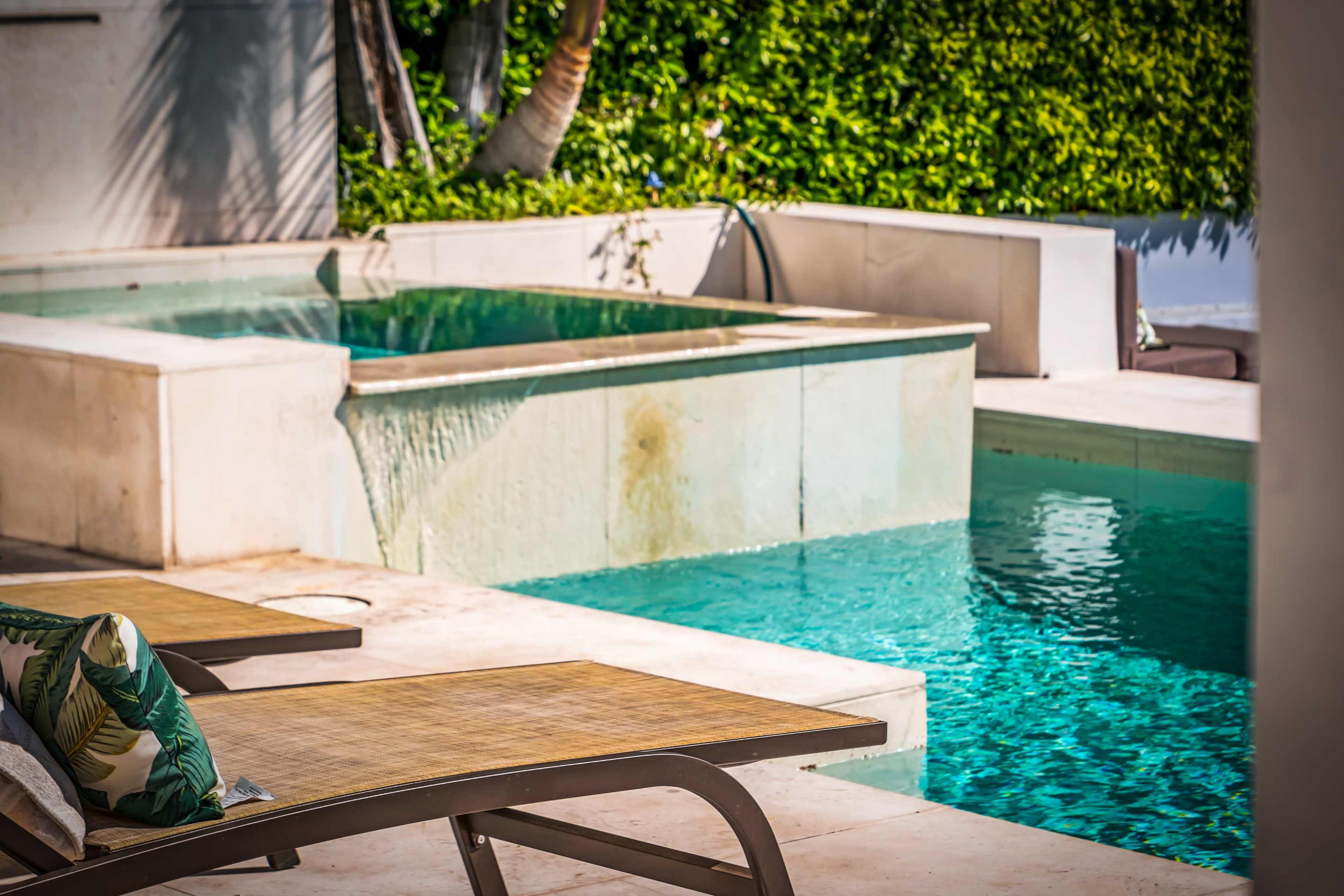The image shows a sunny outdoor area featuring two swimming pools surrounded by lounge chairs and lush greenery.