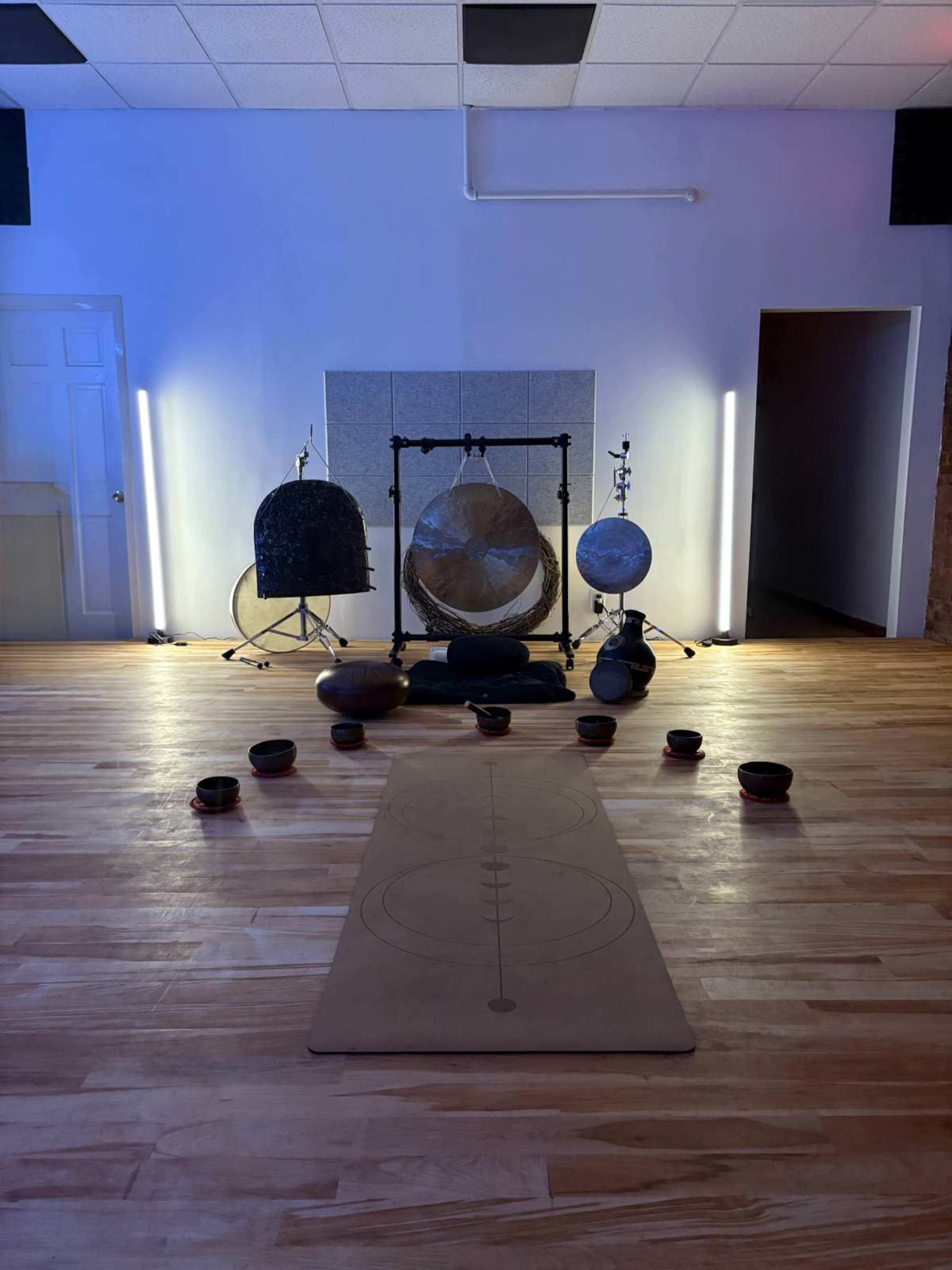 The image shows a meditation space with a mat in the center, surrounded by various sound bowls and instruments, including a gong and a large globe, under soft lighting.