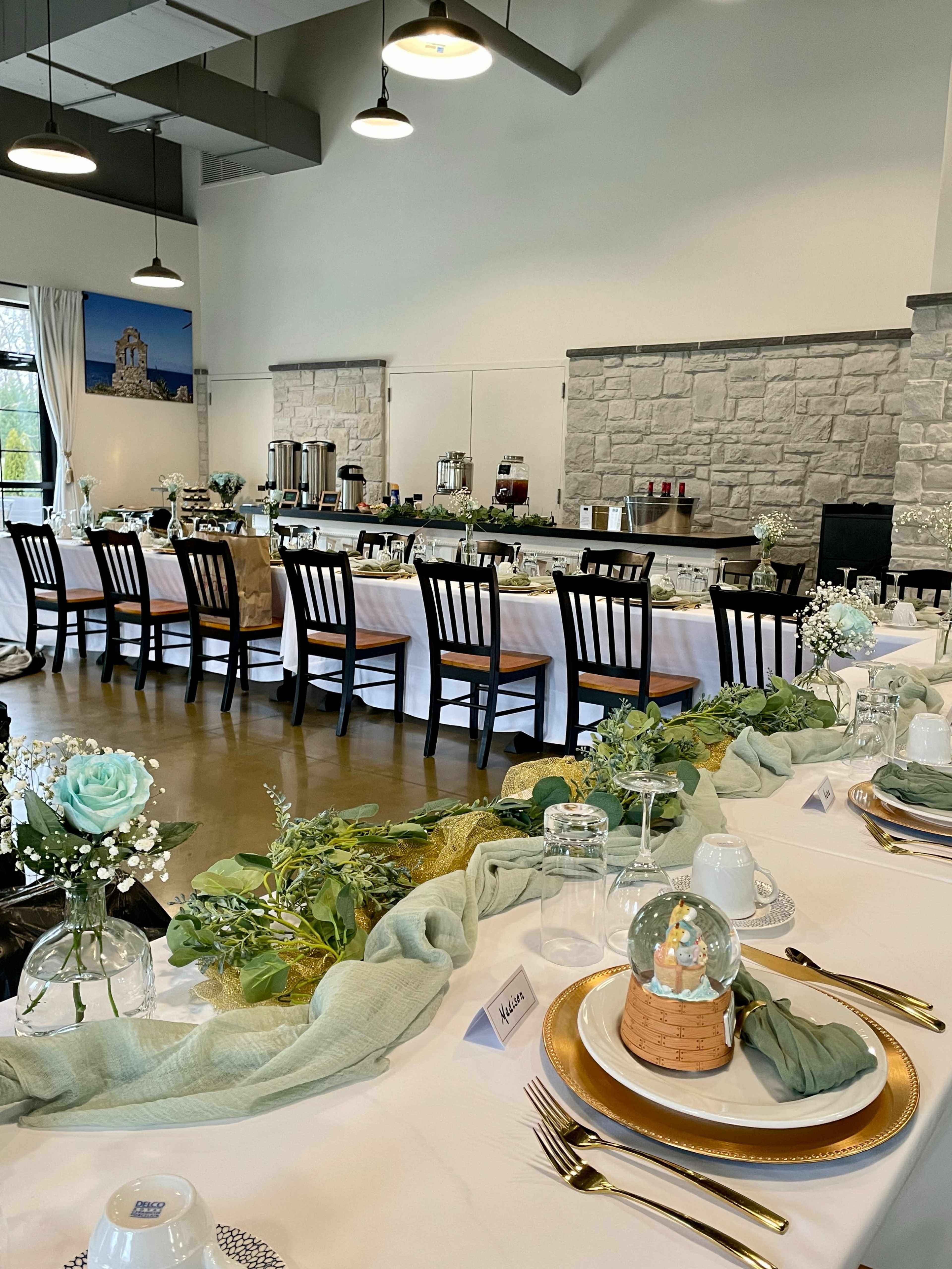 A long dining table set for an event features elegant table decorations, including flowers and greenery, in a well-lit venue with stone walls.