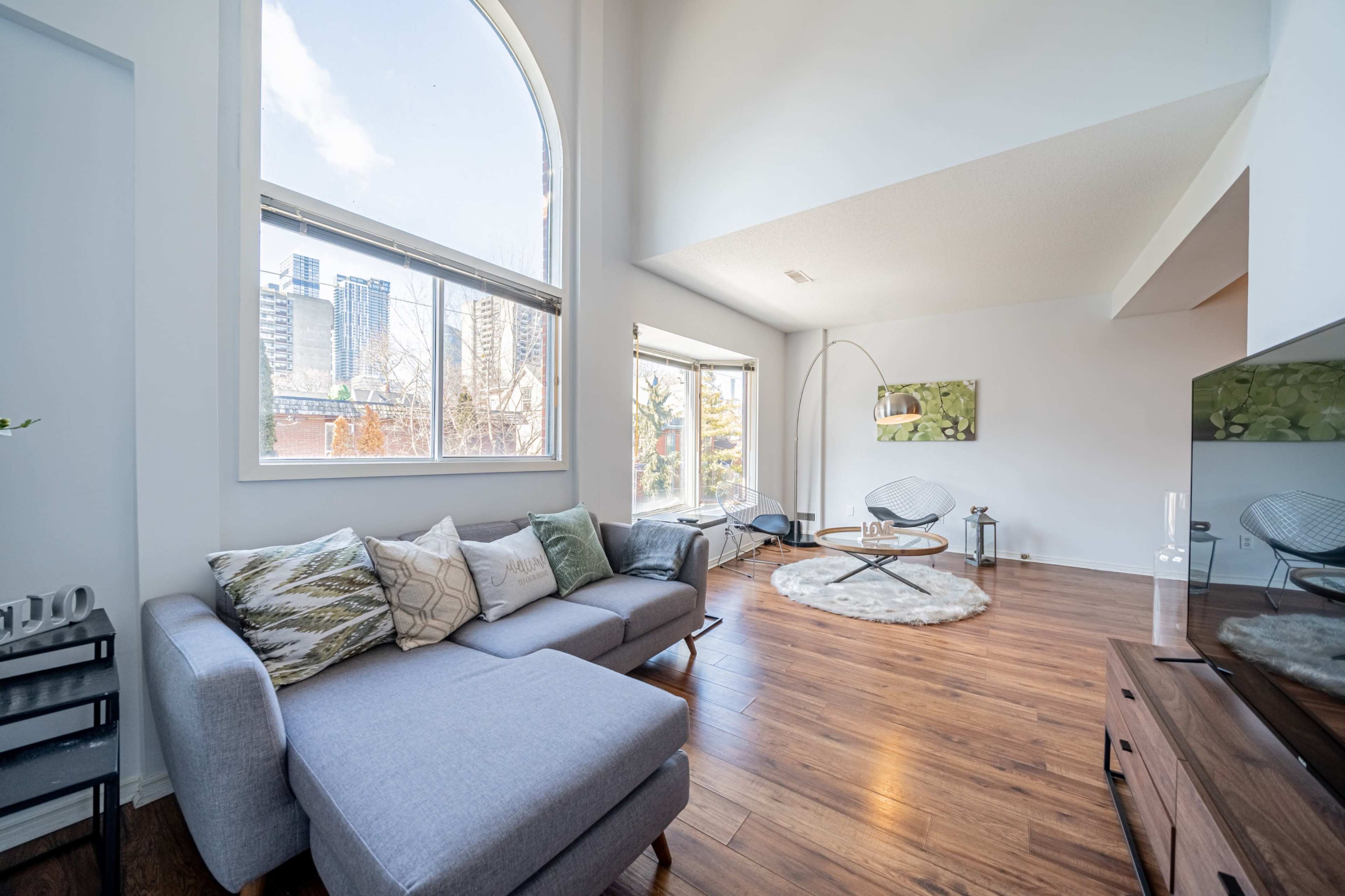 A modern living room features a gray sectional sofa, a round rug on wooden flooring, and large windows showcasing a city view.