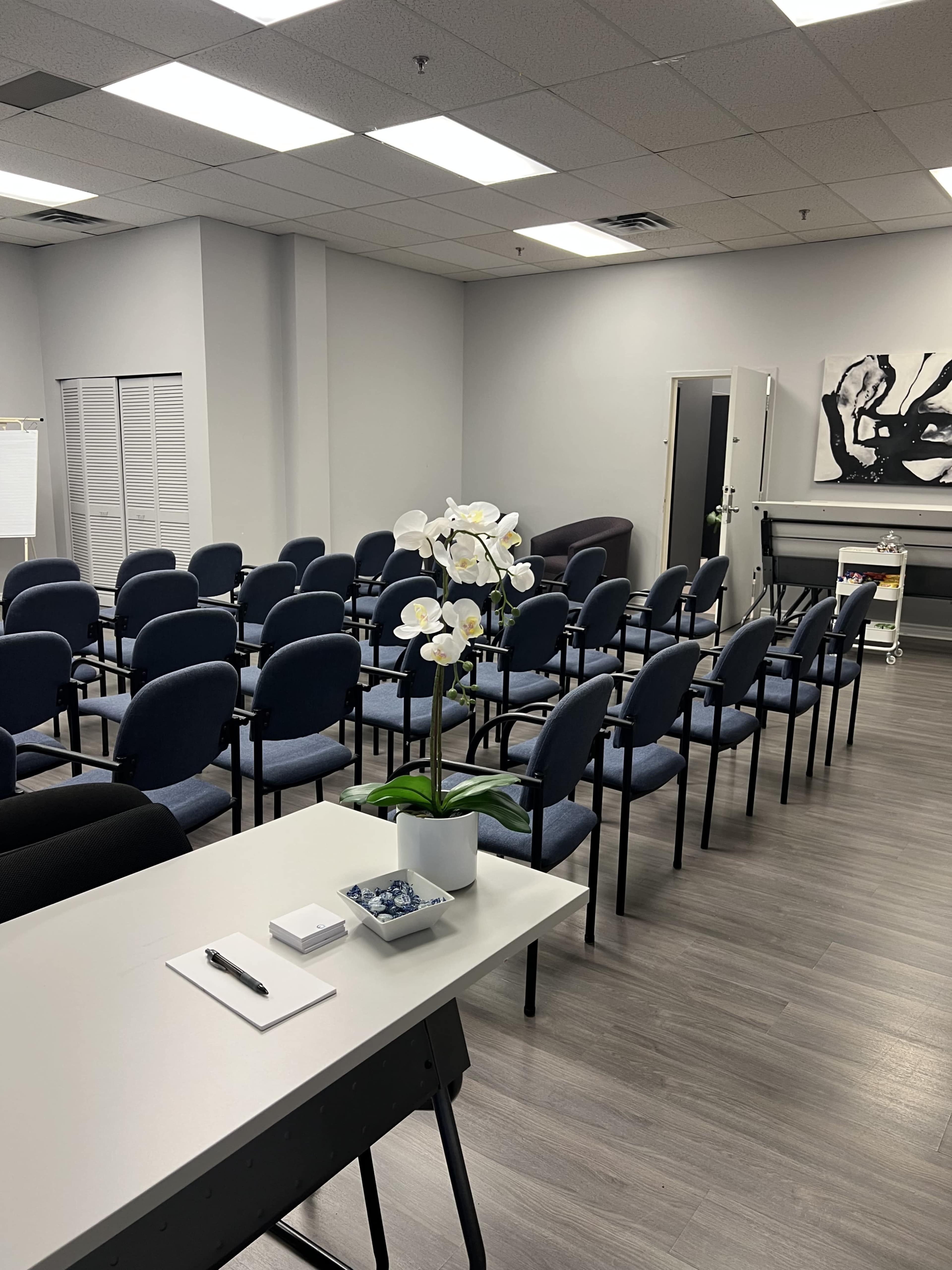 The image shows a meeting room with rows of blue chairs facing a wall, a table with an orchid plant in the foreground, and a whiteboard on one side.