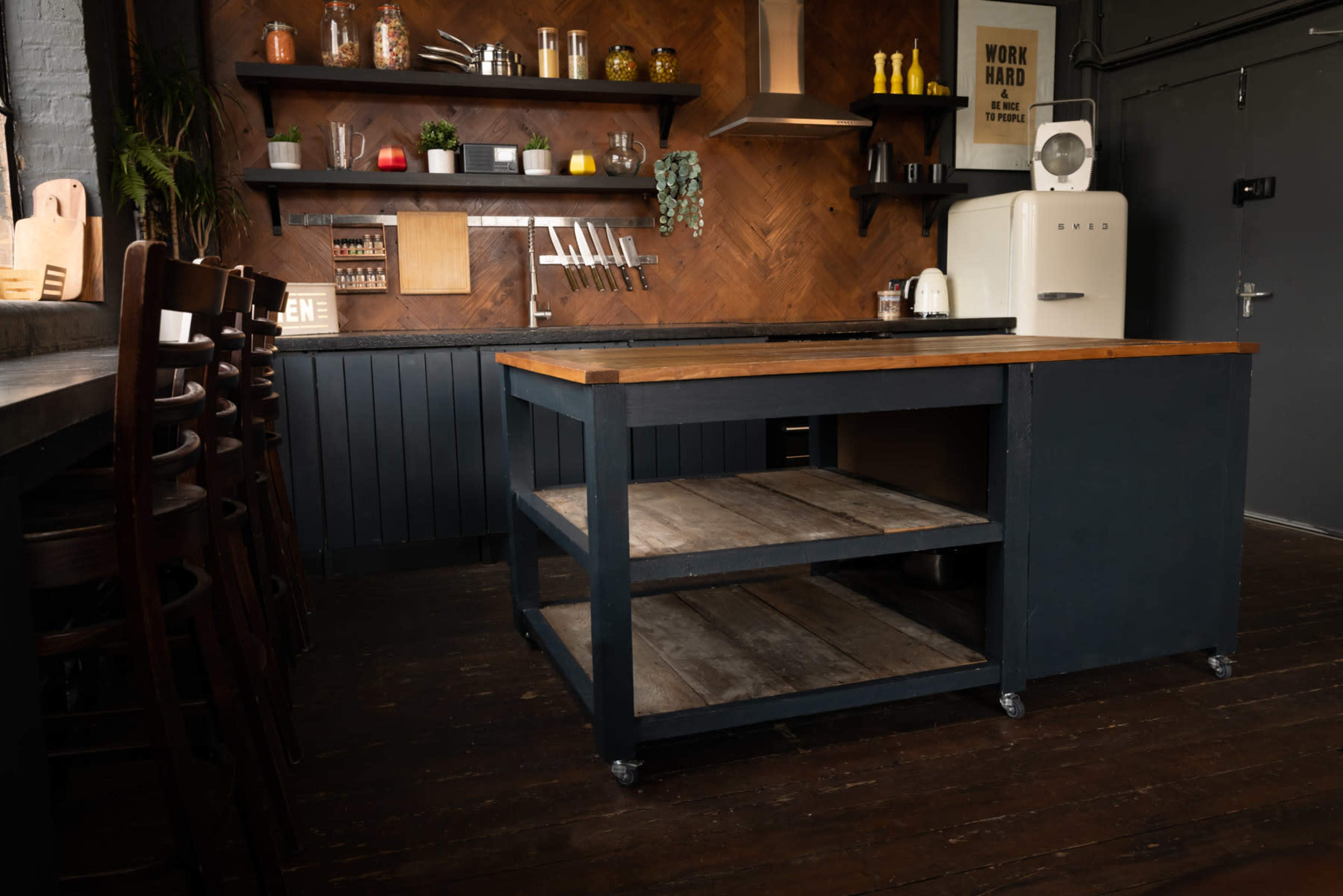A modern kitchen with a dark wooden floor, an island with a wooden top and shelves, and a wall shelf displaying kitchenware and plants.