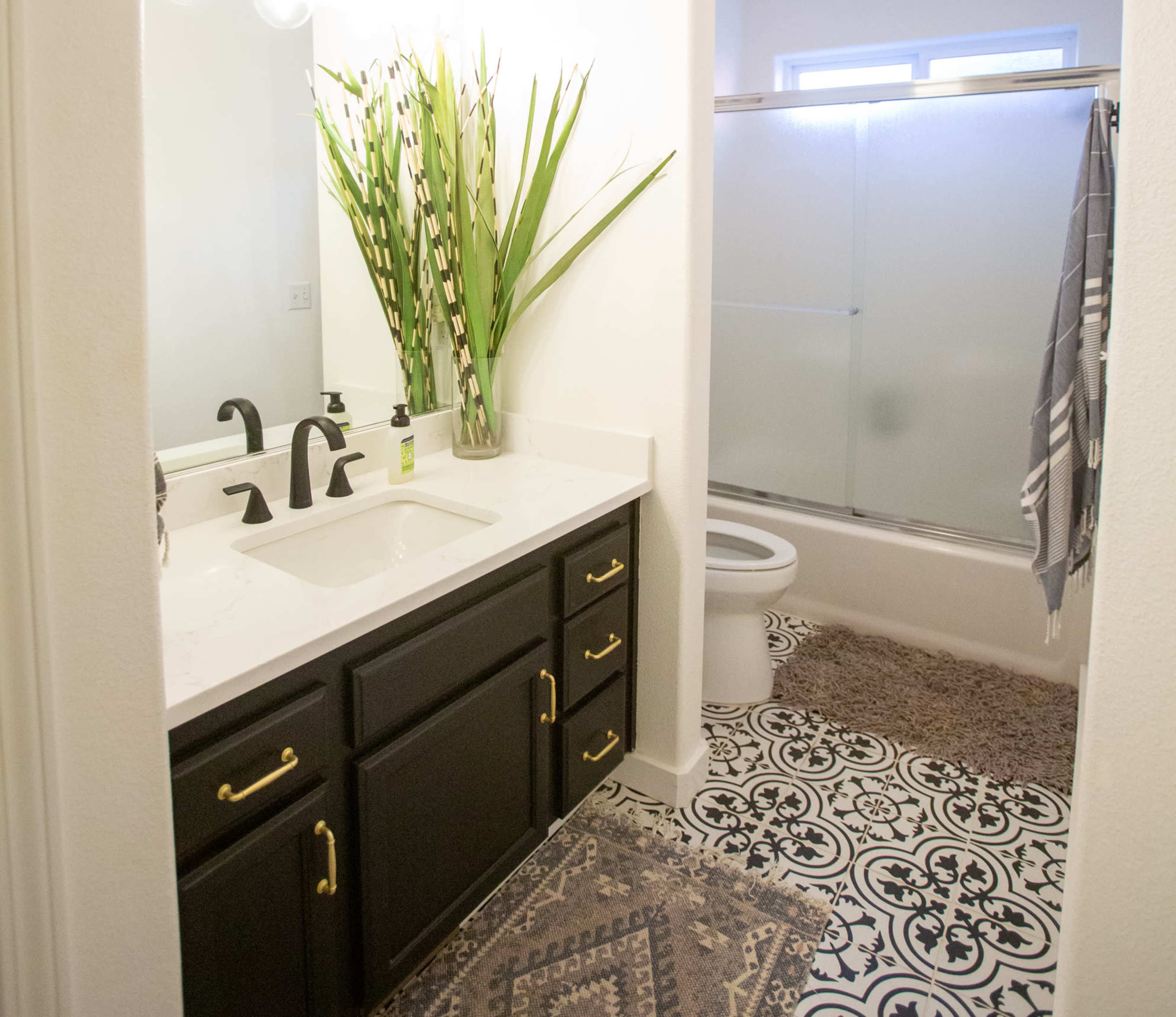 A bathroom featuring a dark wooden vanity with a sink, a large potted plant, a shower with a frosted glass door, and patterned tile flooring.