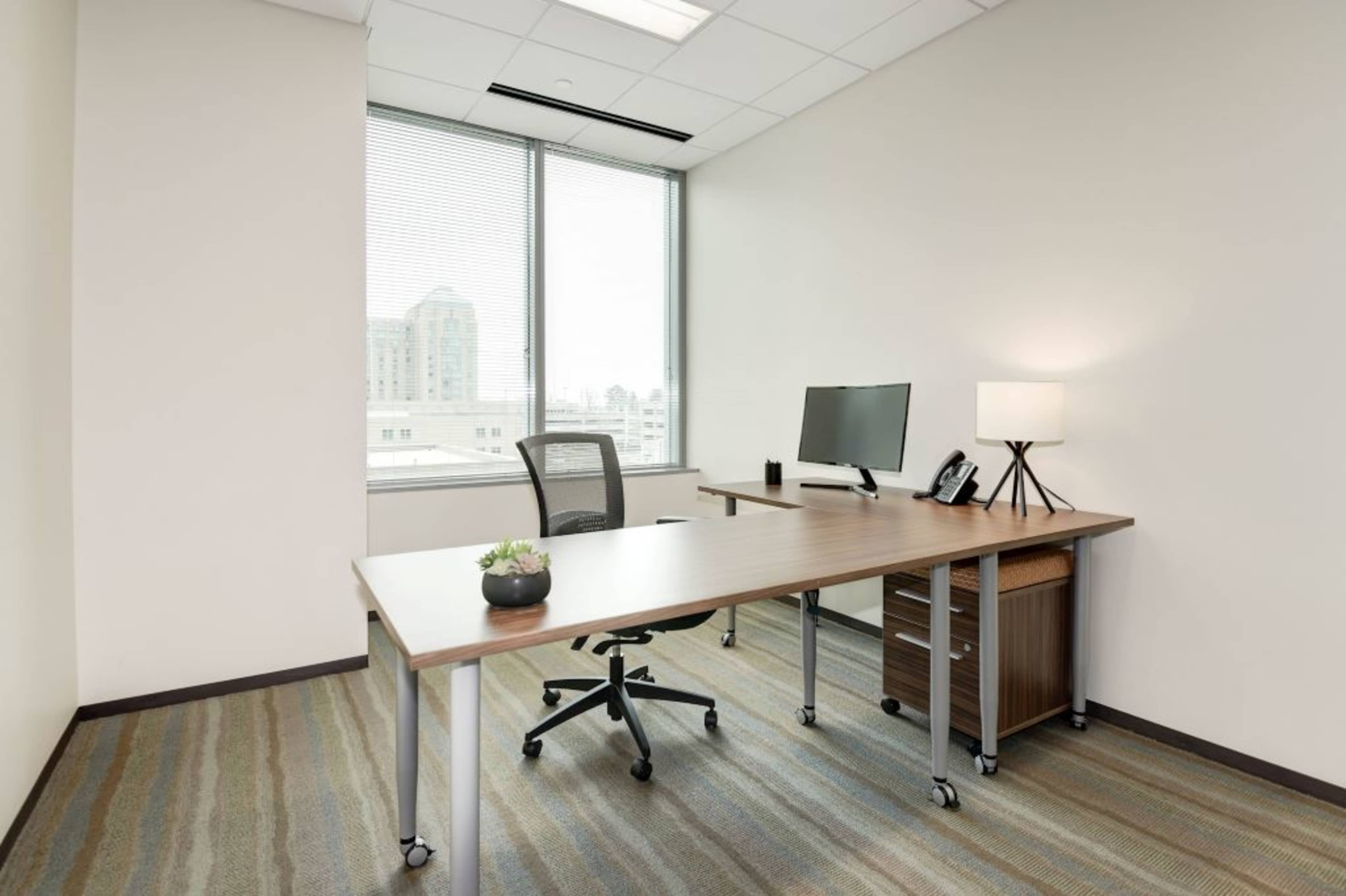 A modern office space featuring a wooden desk, an ergonomic chair, a computer monitor, a lamp, and a small plant on the desk, with large windows offering a view of a cityscape.