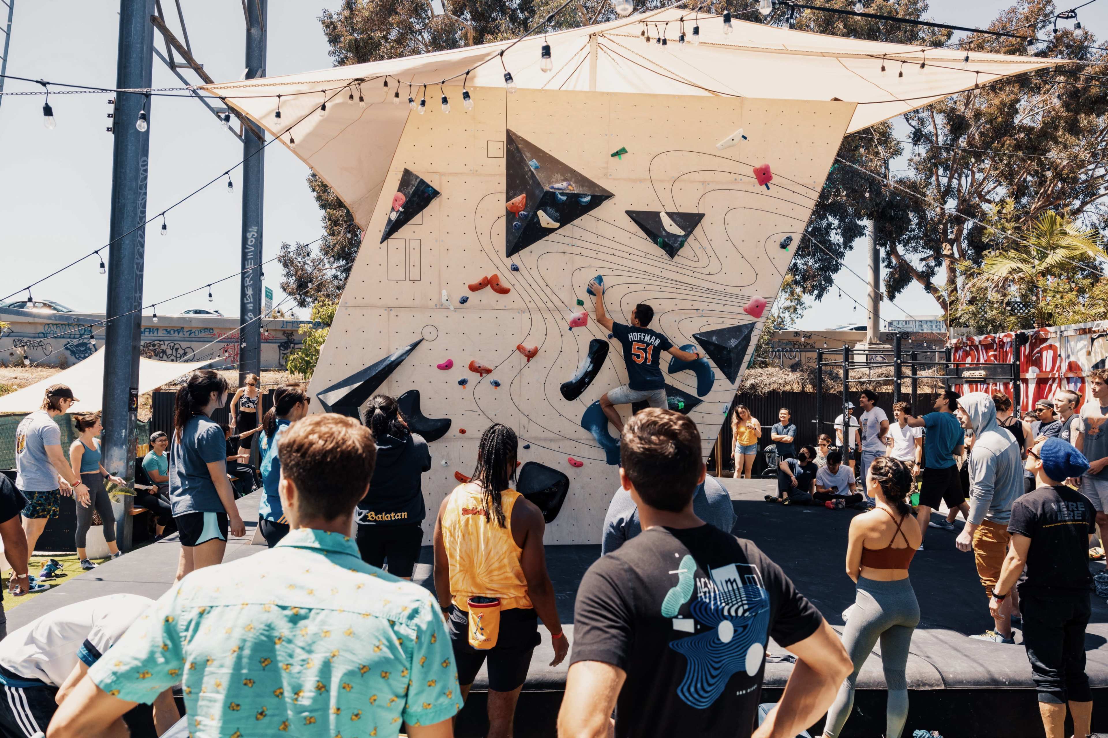 A group of individuals watches as a climber scales a bold climbing wall at an outdoor climbing event.