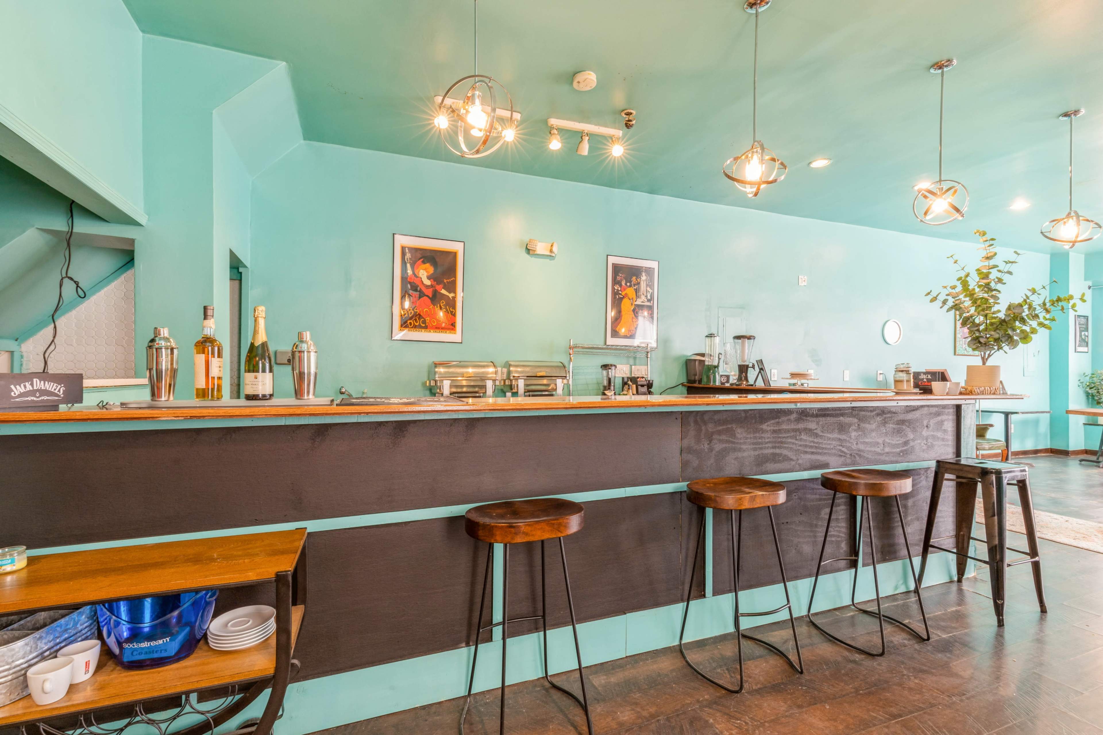 The image shows a modern café interior with a turquoise wall, a dark wooden bar counter, and several metal stools.