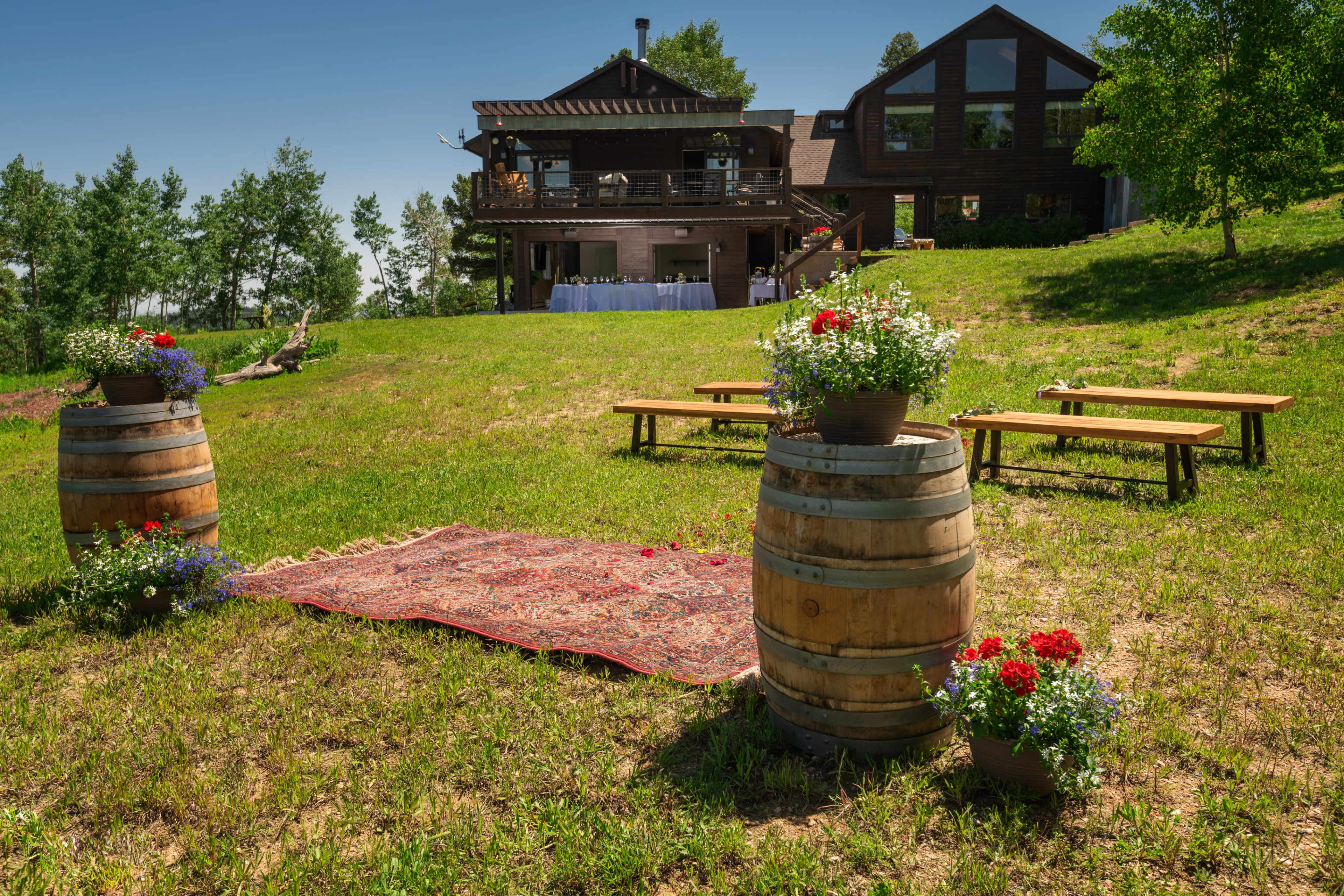 A decorated outdoor setting with wooden barrels, flower pots, and benches is arranged on a grassy lawn in front of a large wooden house.