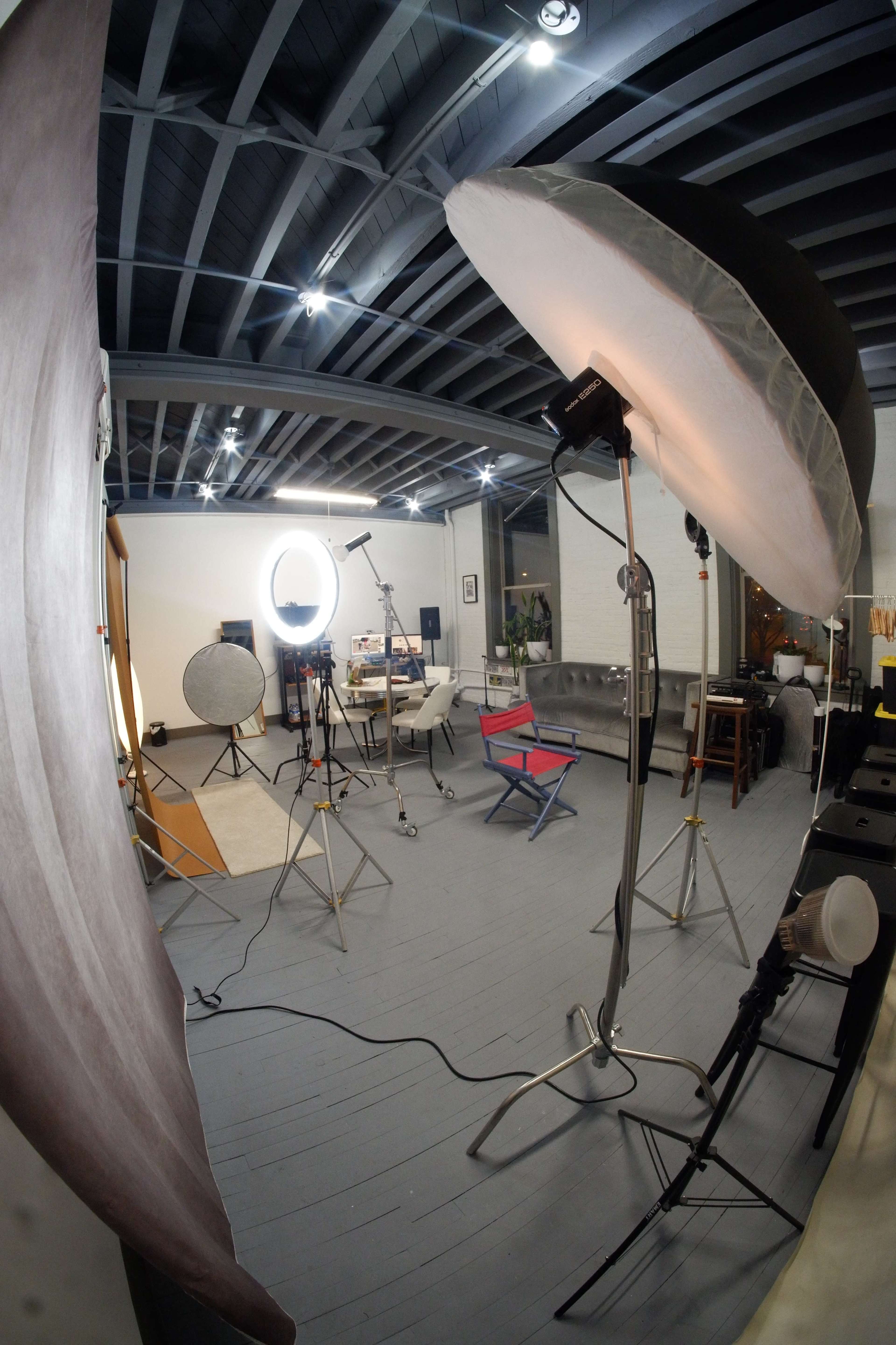 The image shows a photography studio setup with various lighting equipment, a backdrop, and a minimalist seating area.