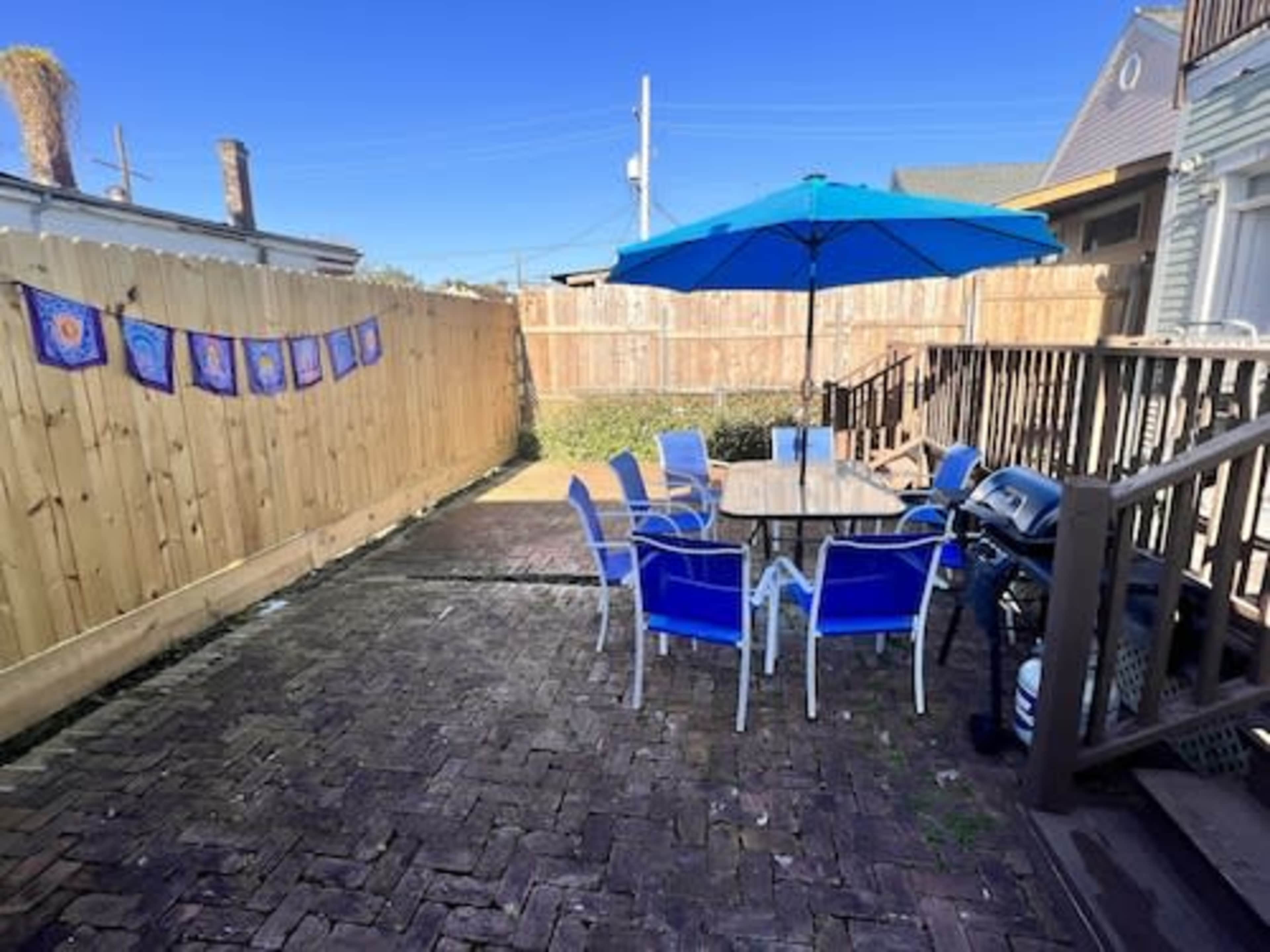 A patio area features a table with eight blue chairs, an umbrella, and a barbecue grill, surrounded by wooden fencing and a clear blue sky.