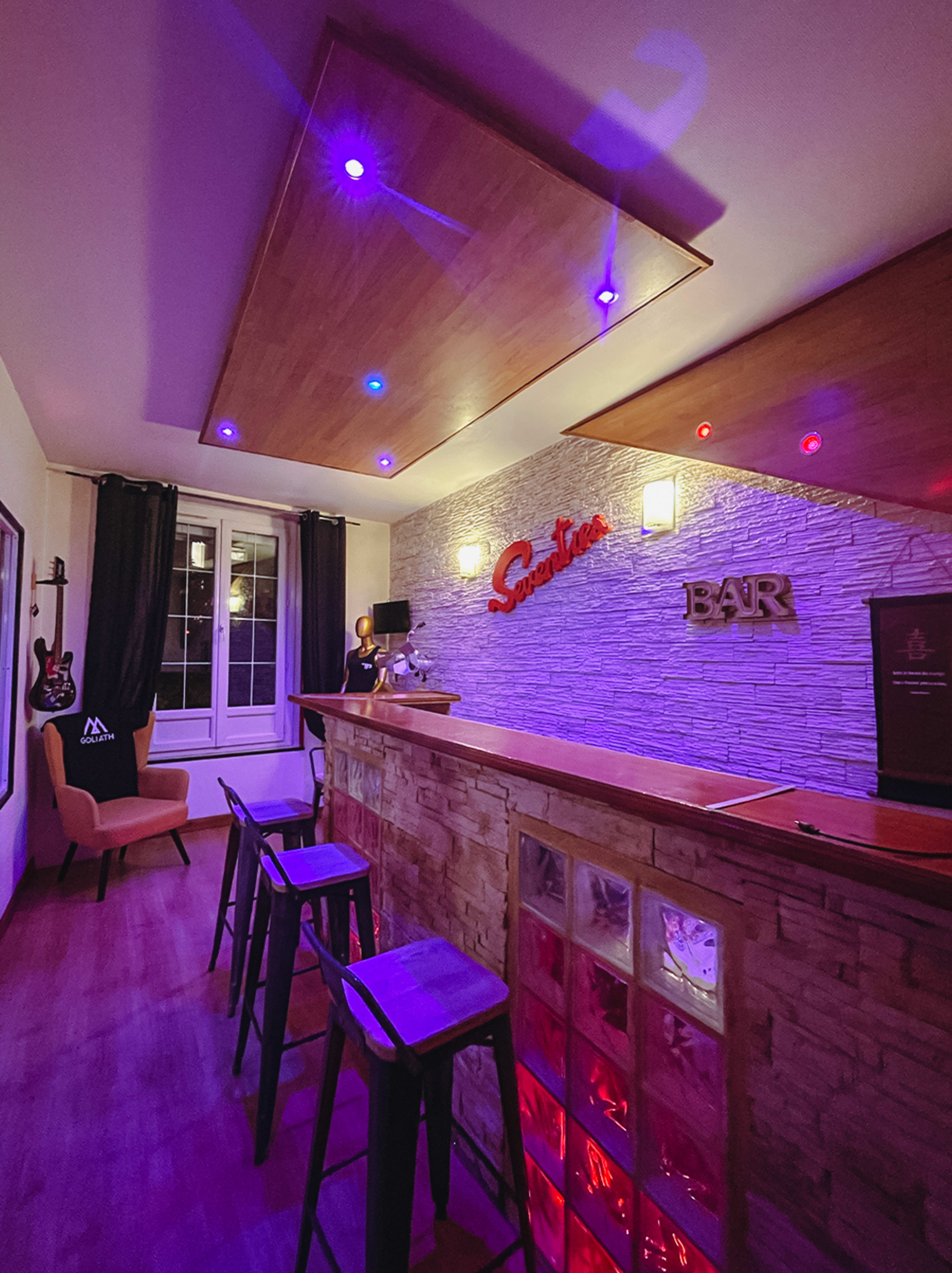 The image shows a modern bar area with a stone accent wall, illuminated by colorful LED lights, featuring bar stools and a seating area.