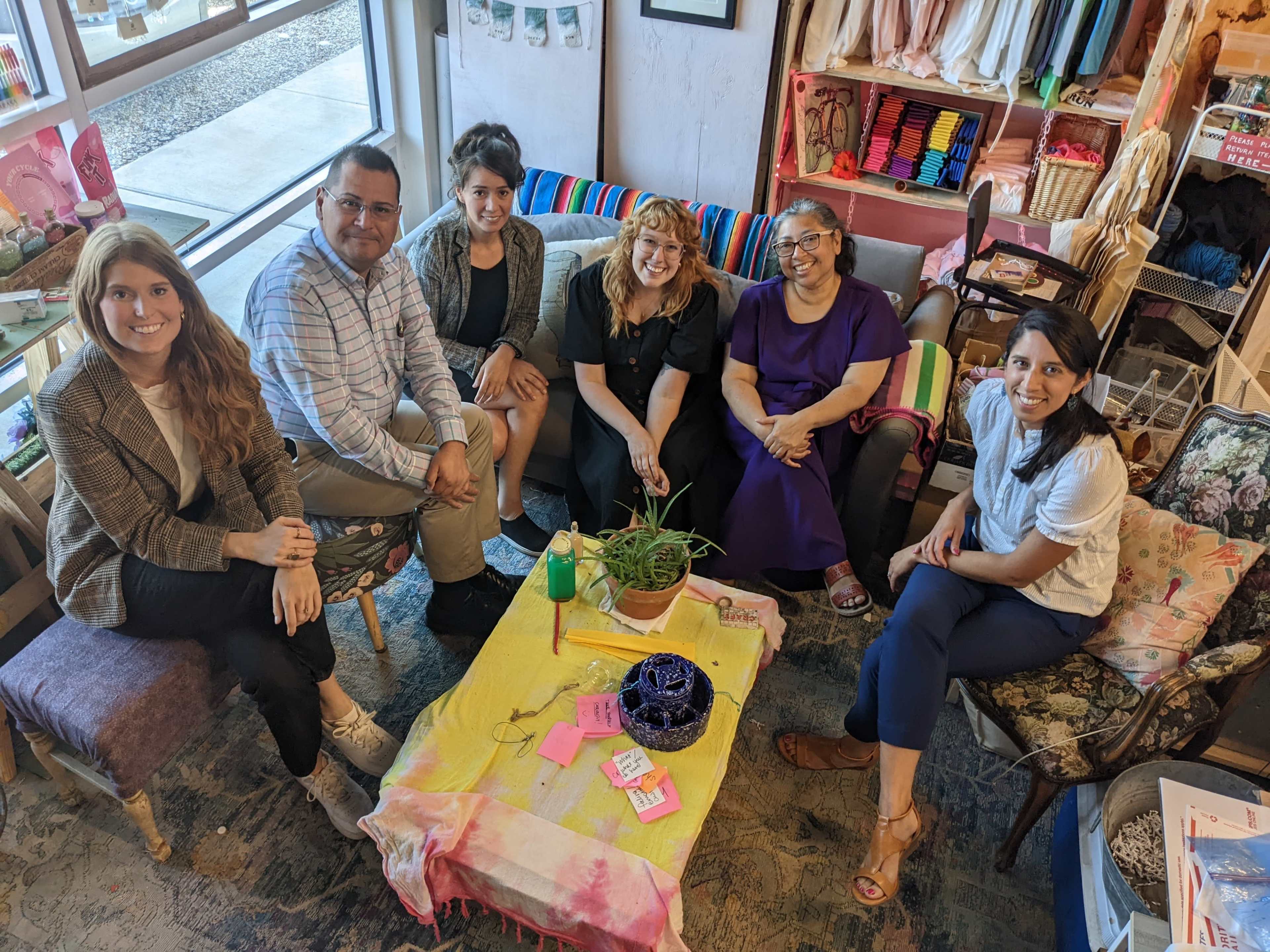 A group of six people sit around a small table in a cozy room filled with colorful fabric and craft supplies.