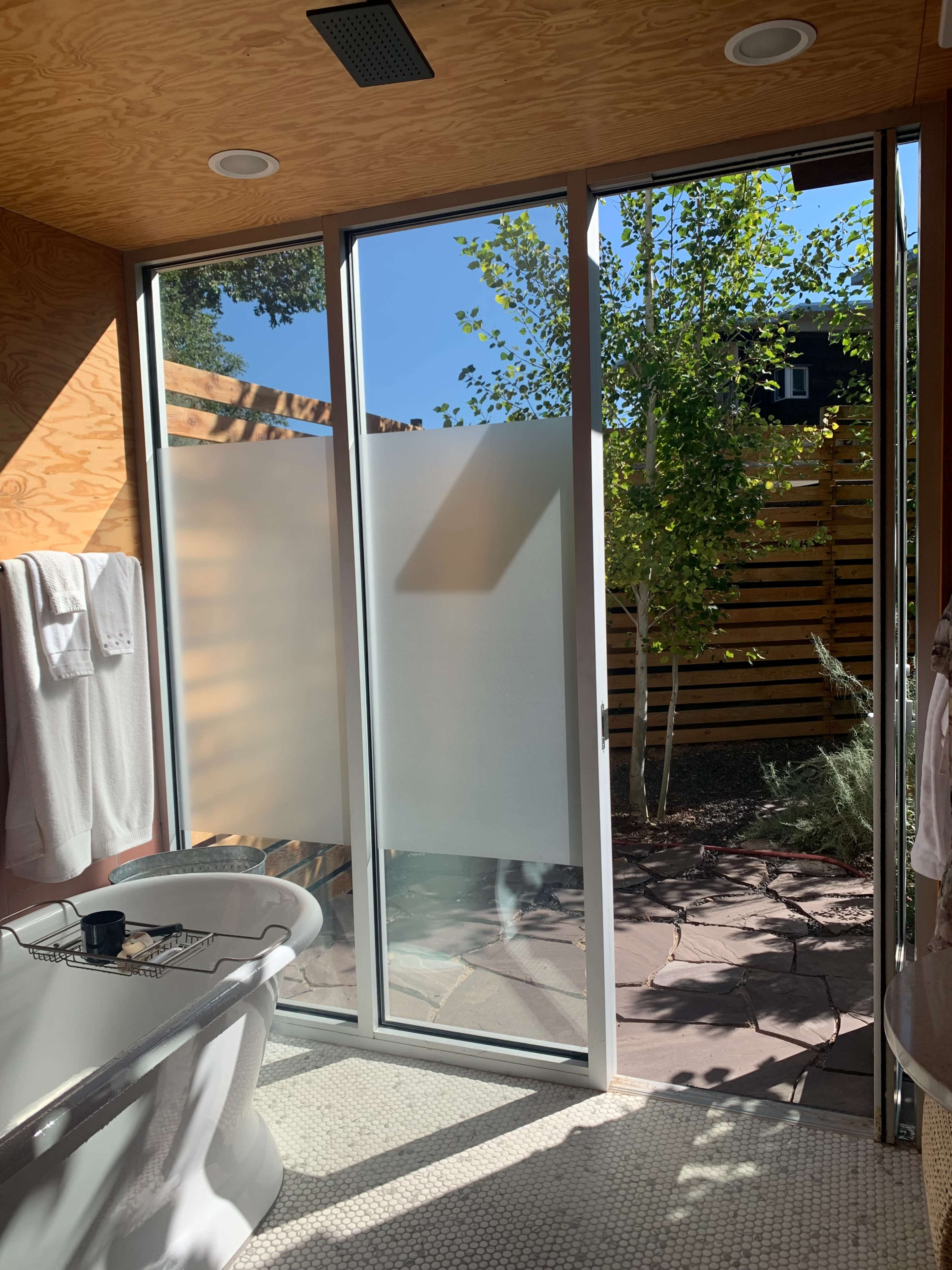 A modern bathroom features a freestanding bathtub next to large glass doors leading to an outdoor space with plants.