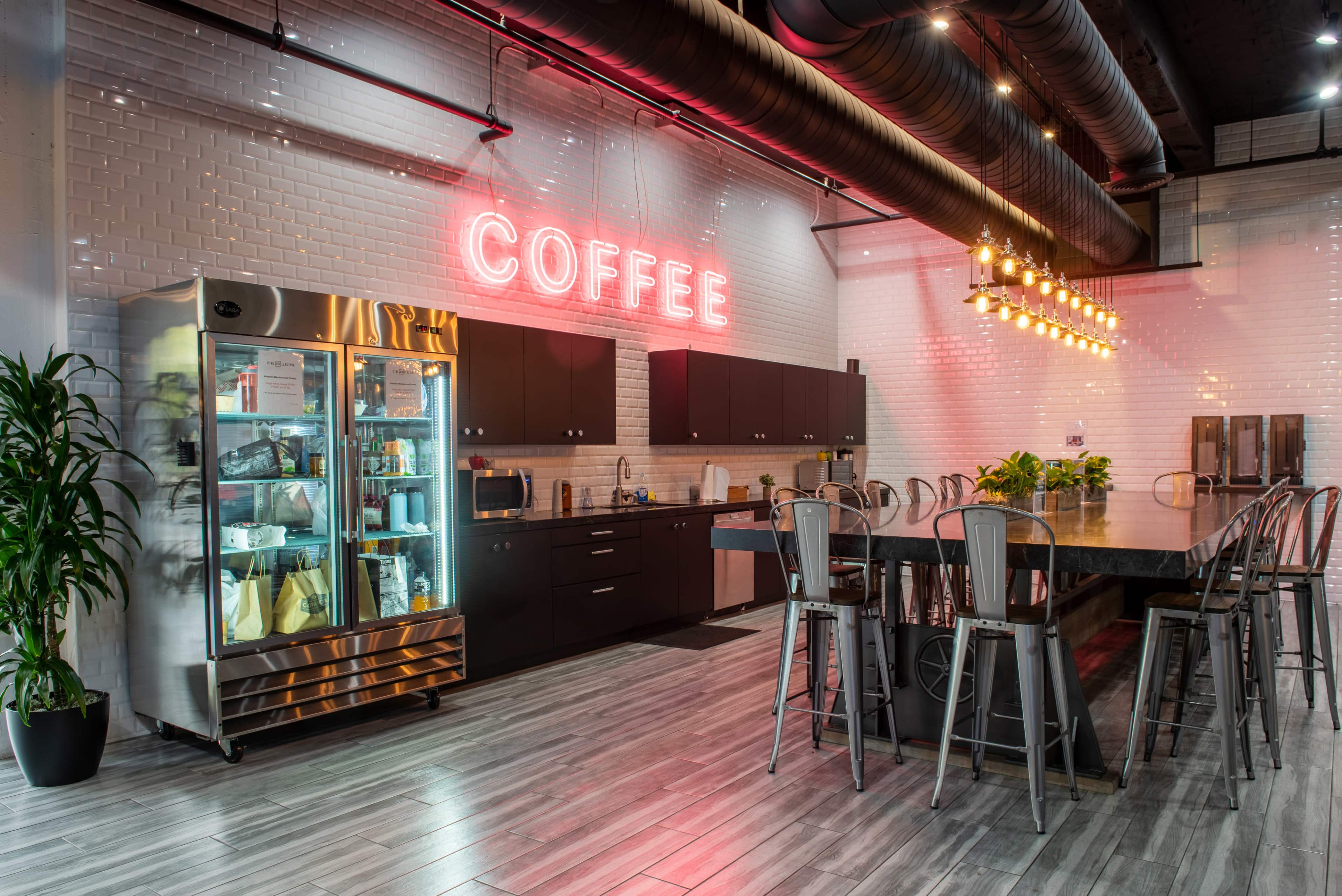 A modern kitchen space features a large communal table, a refrigerator, and a bright neon sign that reads "COFFEE."