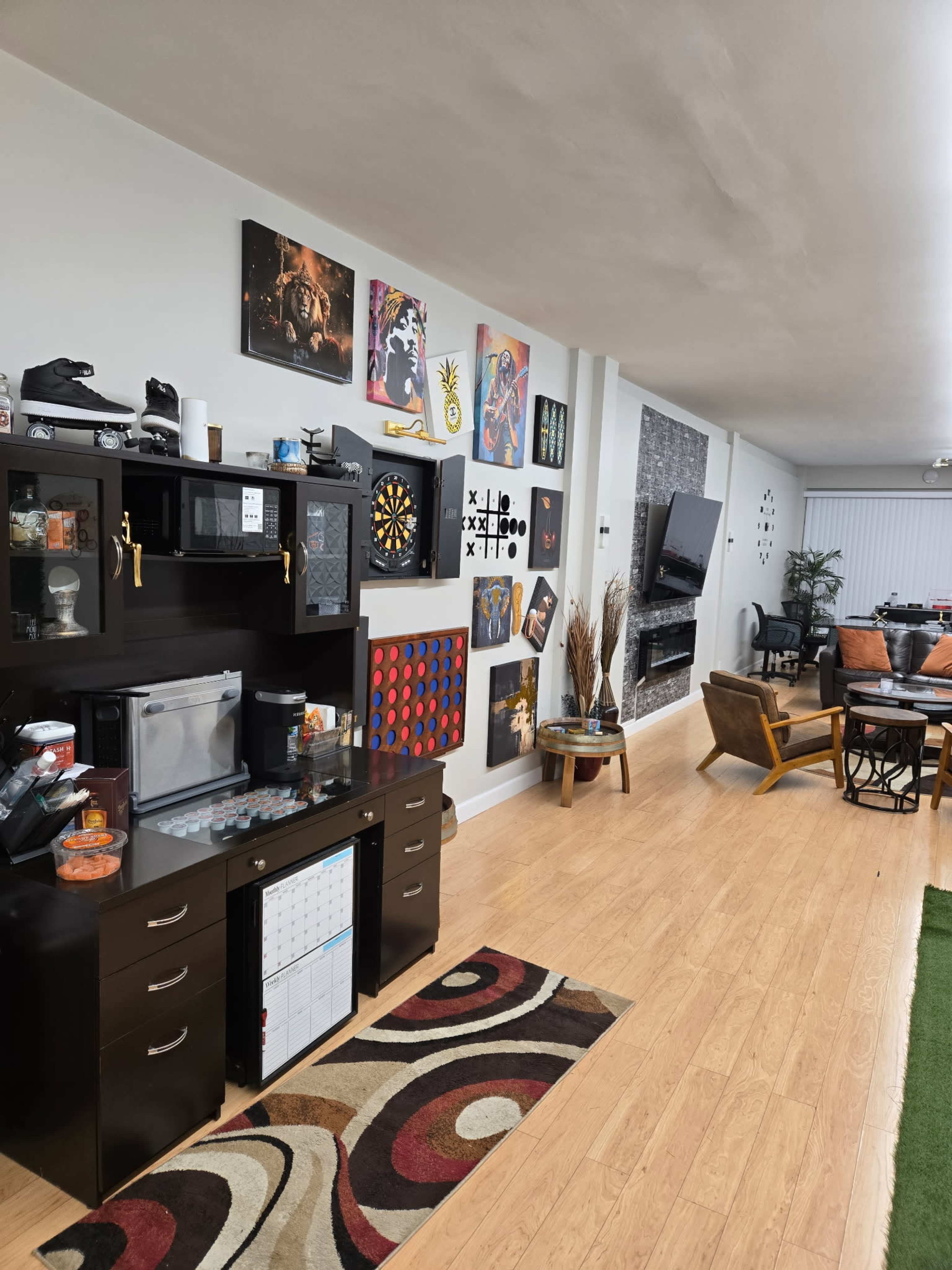 The image shows a living space with a wooden floor, featuring a wall adorned with various framed artworks and a small bar area on the left with a refrigerator and storage drawers.