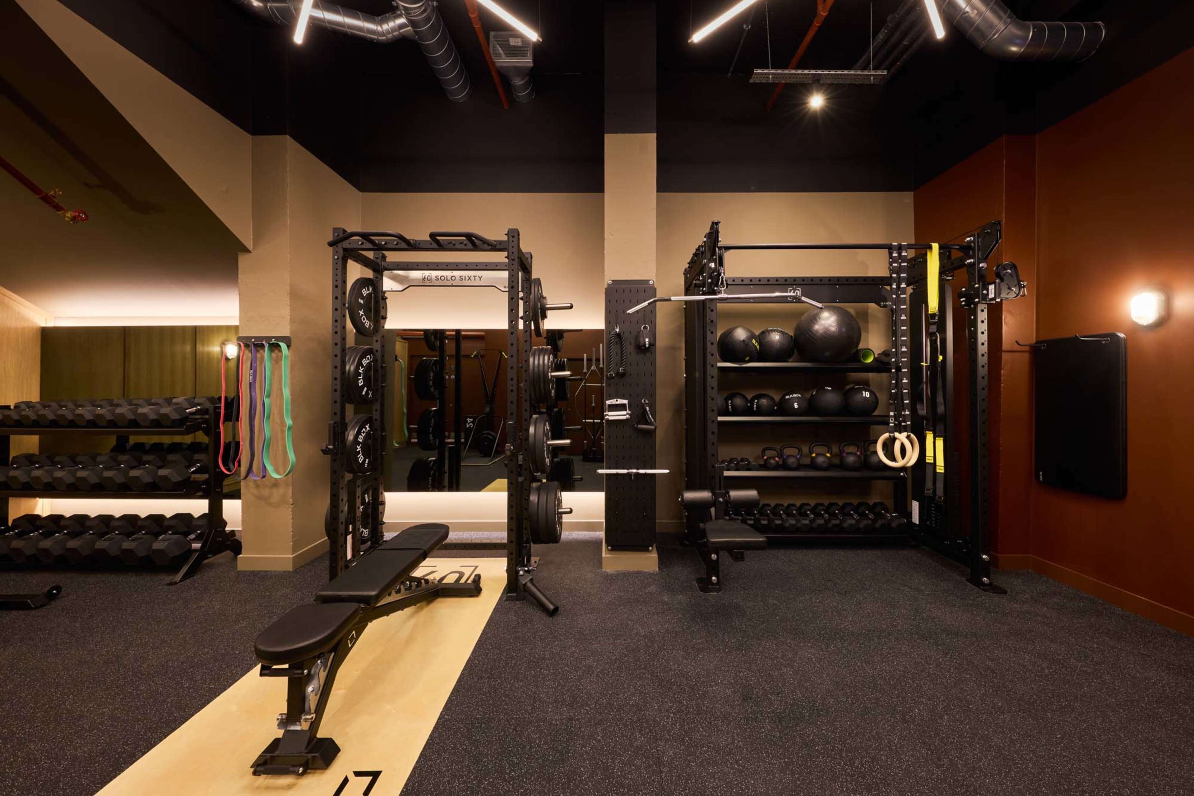The image shows a modern gym interior featuring weightlifting equipment, a bench, and storage racks filled with exercise balls and weights.