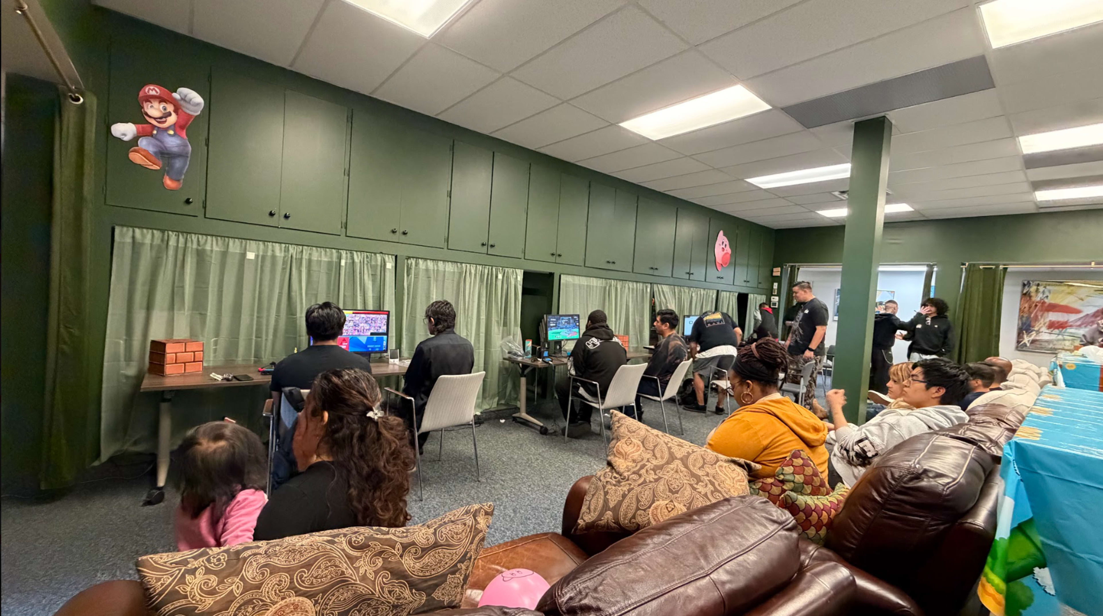 A group of people is seated in a gaming area, with several individuals playing video games at computers, while others watch and socialize in a relaxed environment.