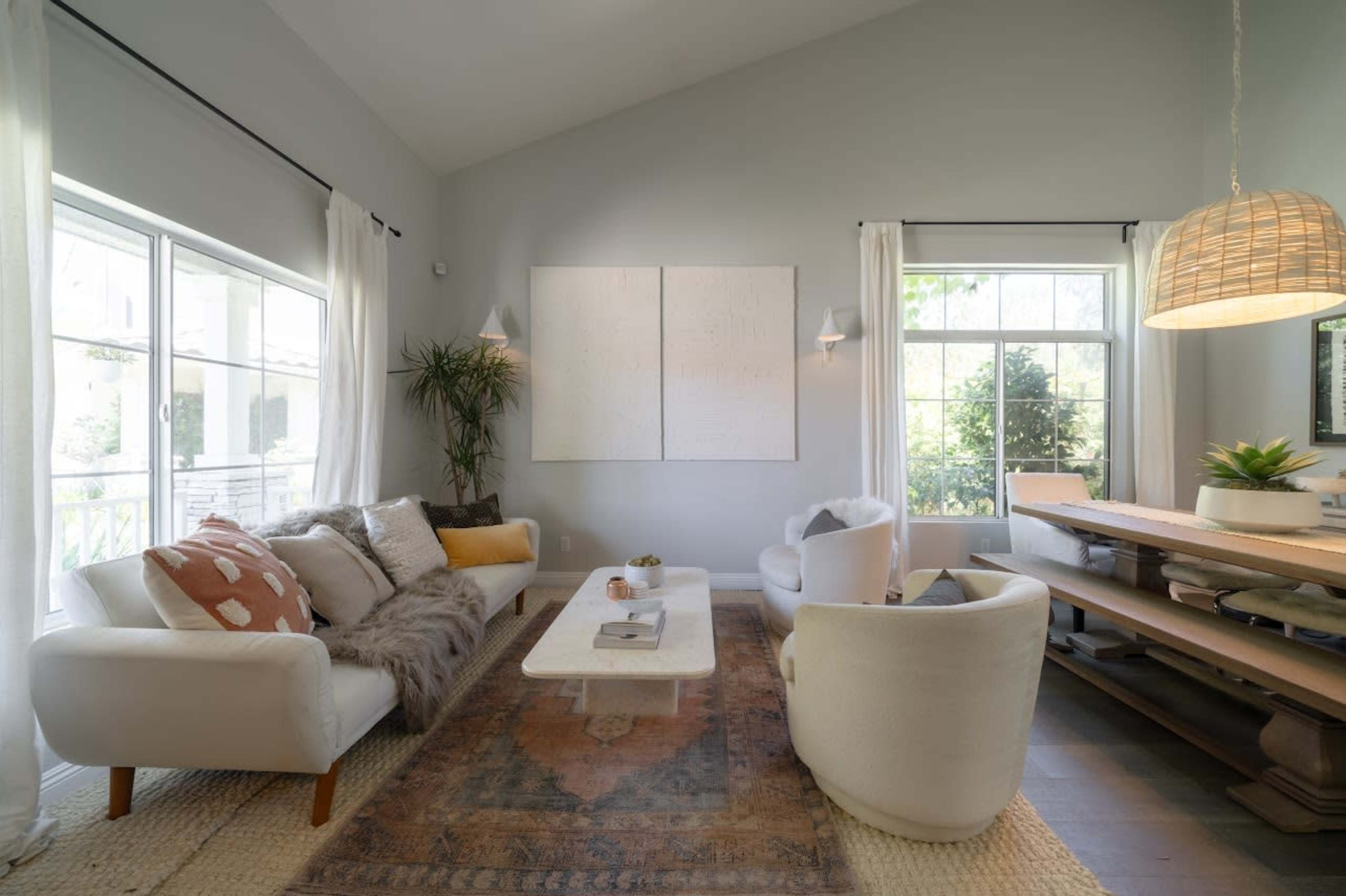 A modern living room features a cozy sofa with patterned cushions, a white coffee table, and a dining area with a wooden table, all complemented by large windows allowing natural light.