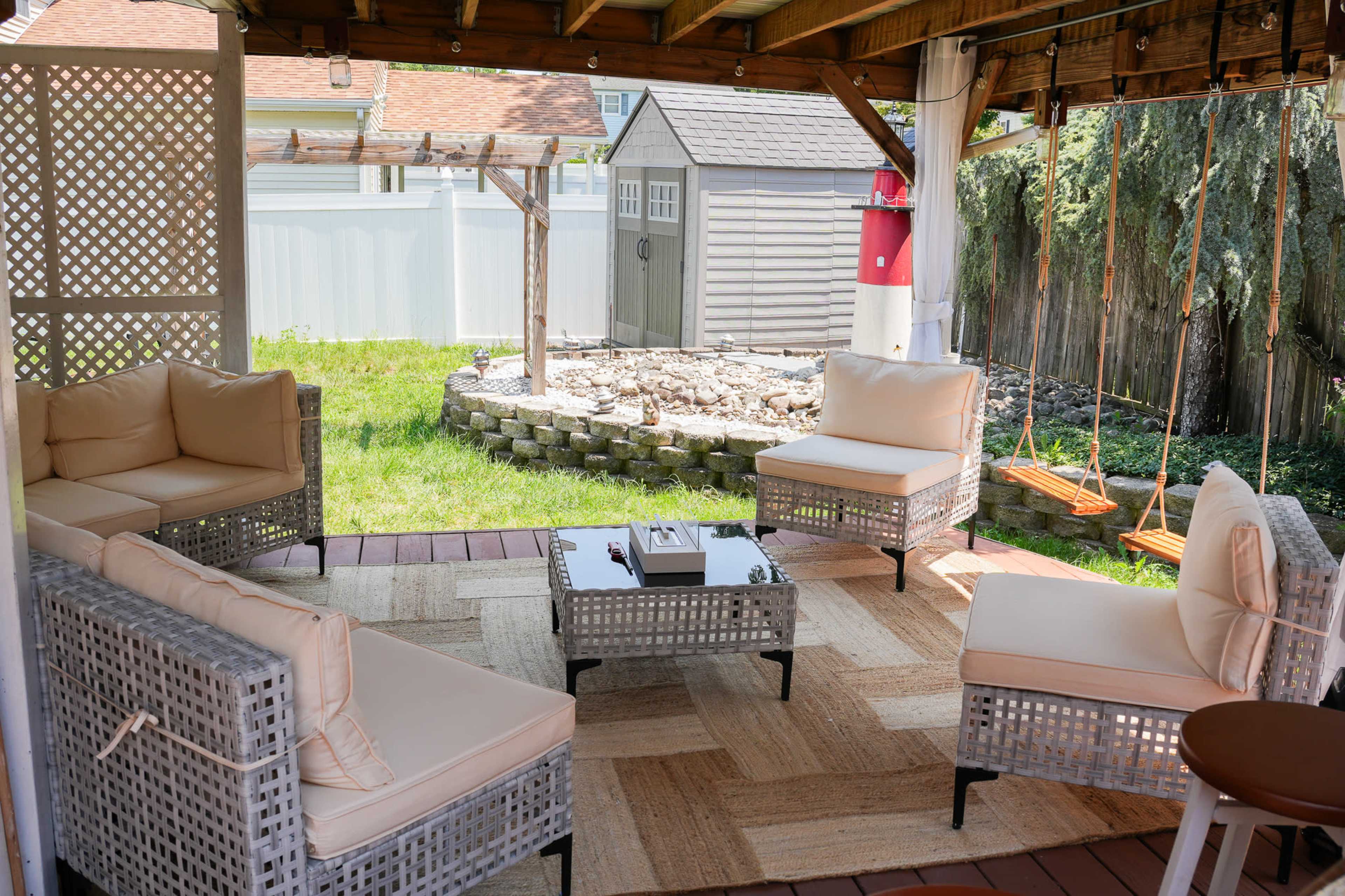 The image shows a cozy outdoor seating area with four cushioned chairs arranged around a small table, a stone perimeter garden in the background, and a shed visible in the distance.