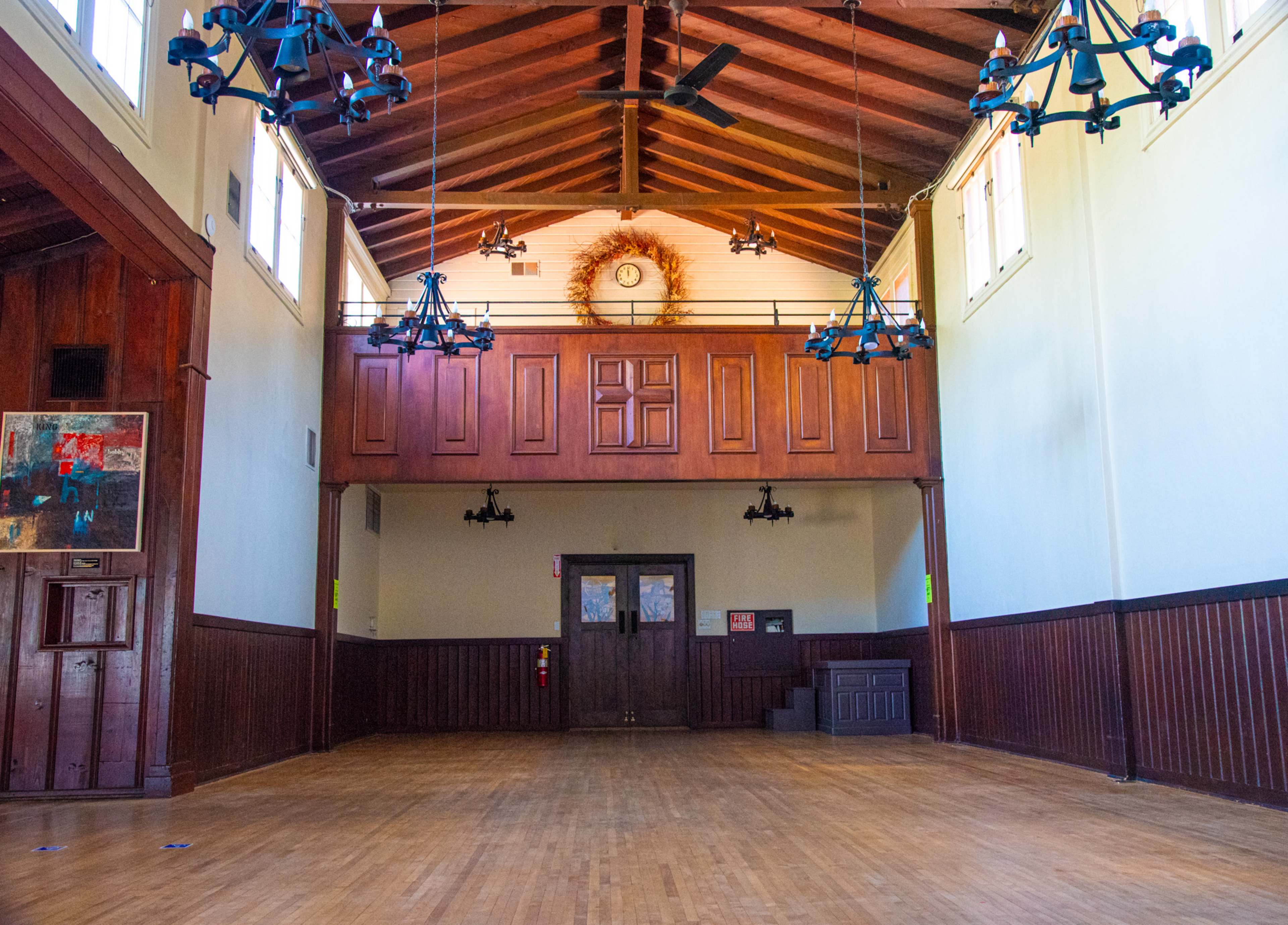 A spacious, wooden-floored hall with high ceilings, featuring wooden paneling, chandeliers, and a balcony at the rear.