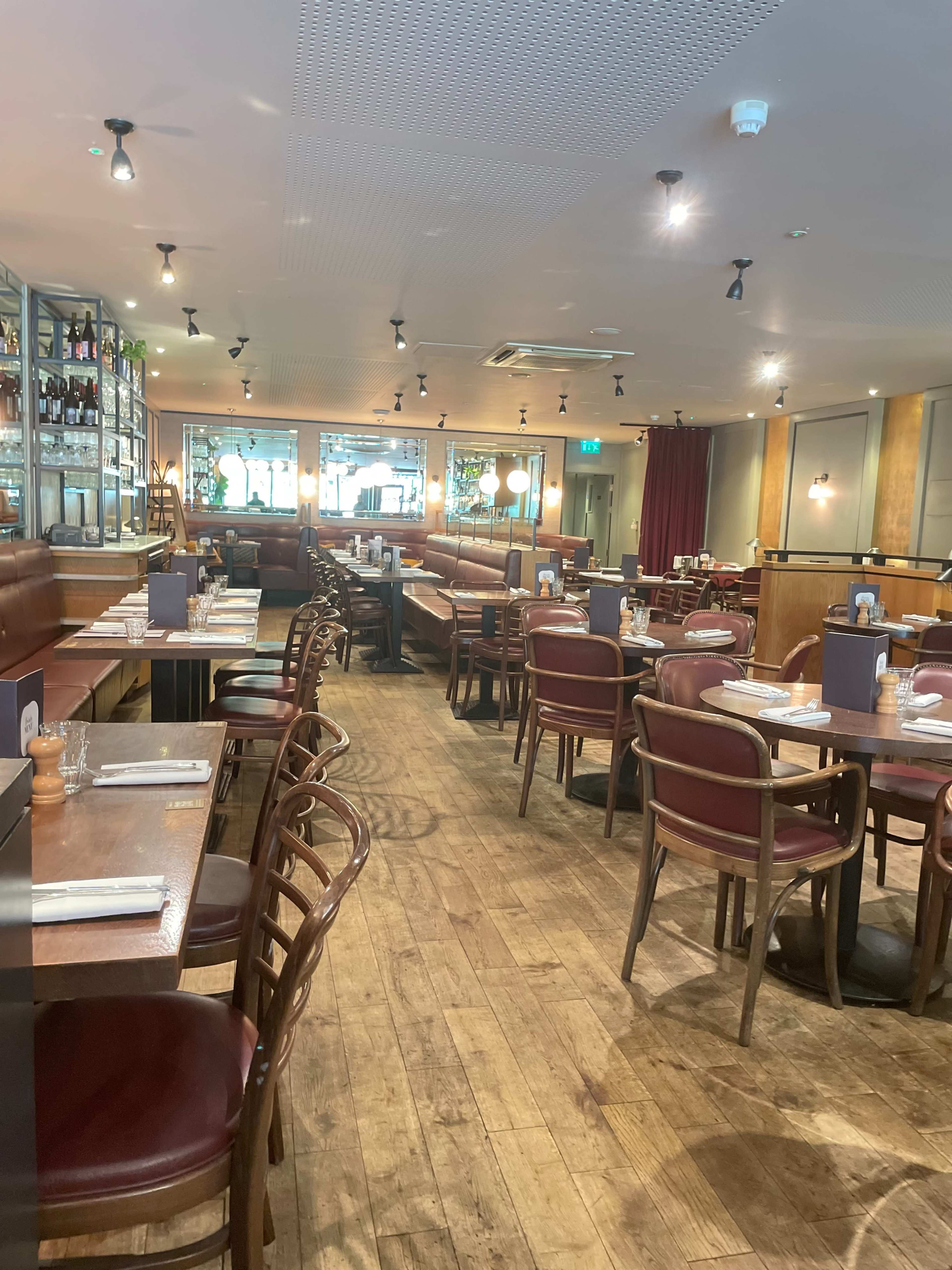 The image shows an empty restaurant interior with wooden floors, rows of tables and chairs, and a bar area in the background.