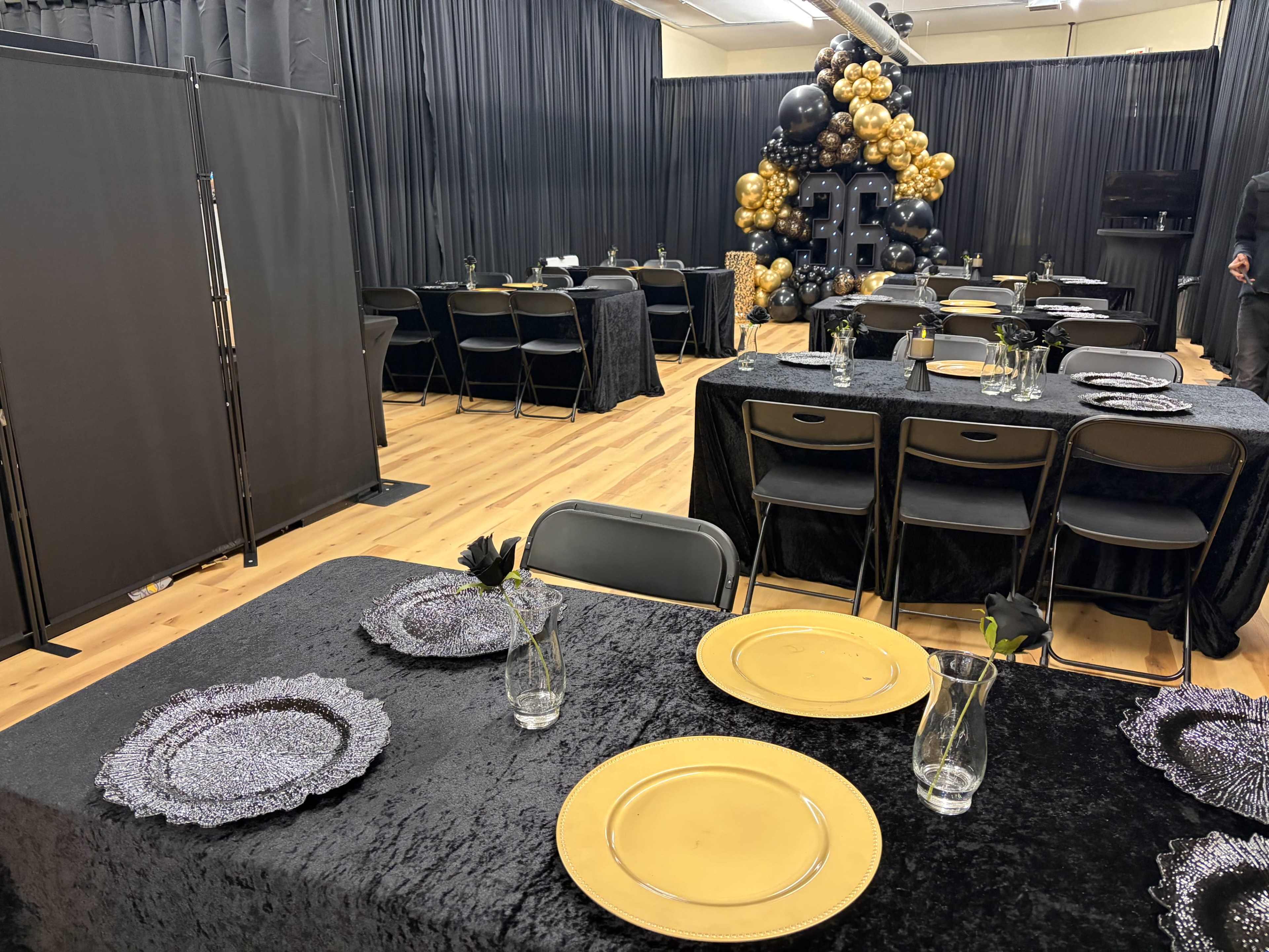 The image shows a decorated event space with tables set with black cloths, gold plates, and a backdrop of black and gold balloons.