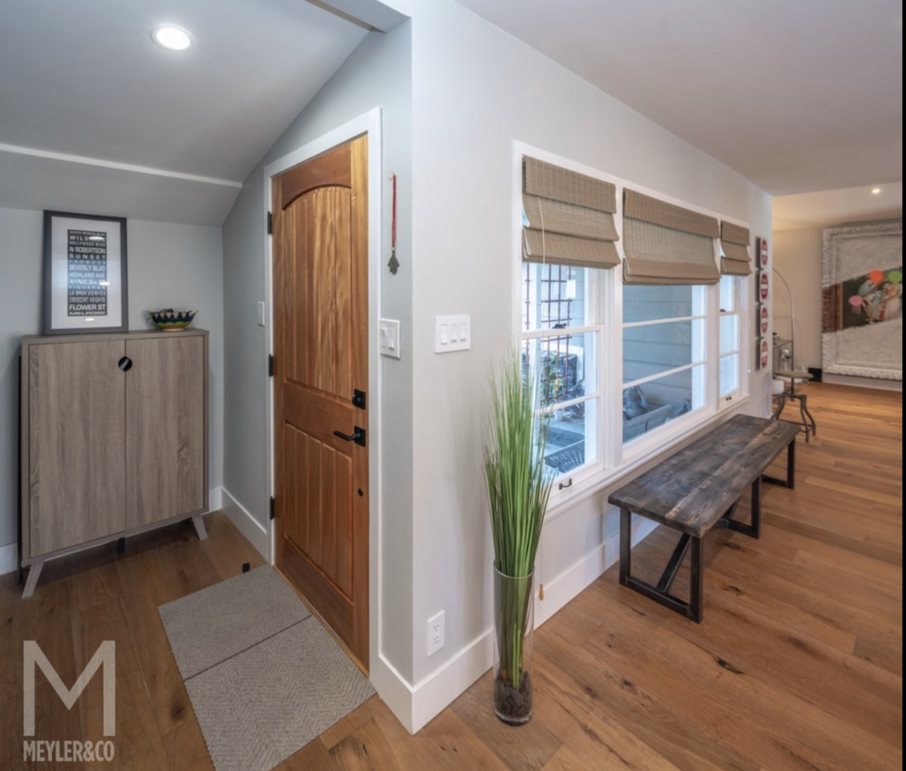 A light-filled entryway with a wooden door, a narrow bench, and a potted plant next to a window, leading into a room with hardwood floors.