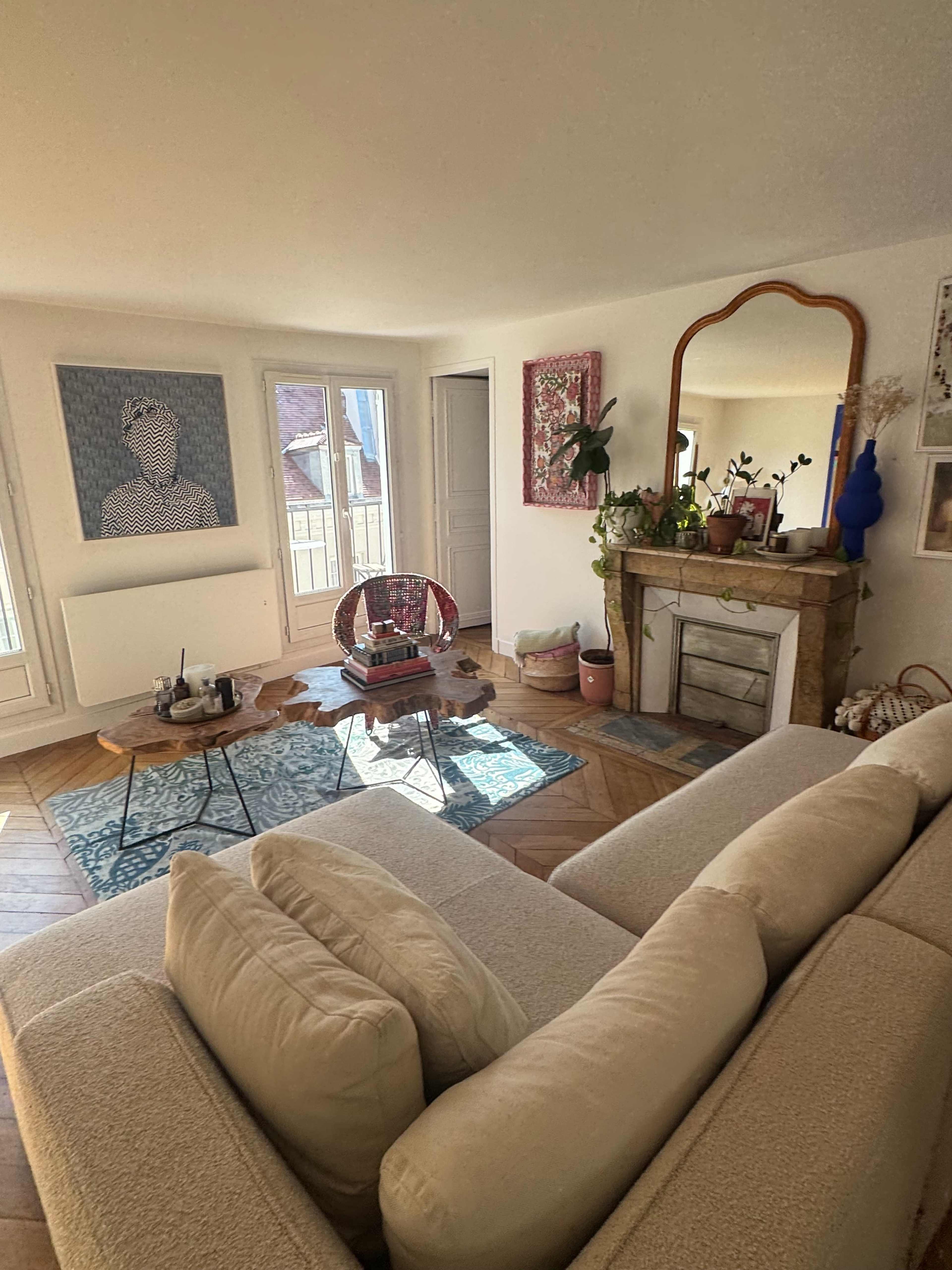 The image shows a bright living room with a beige couch, a coffee table, and a decorative fireplace, along with artwork on the walls and large windows letting in natural light.