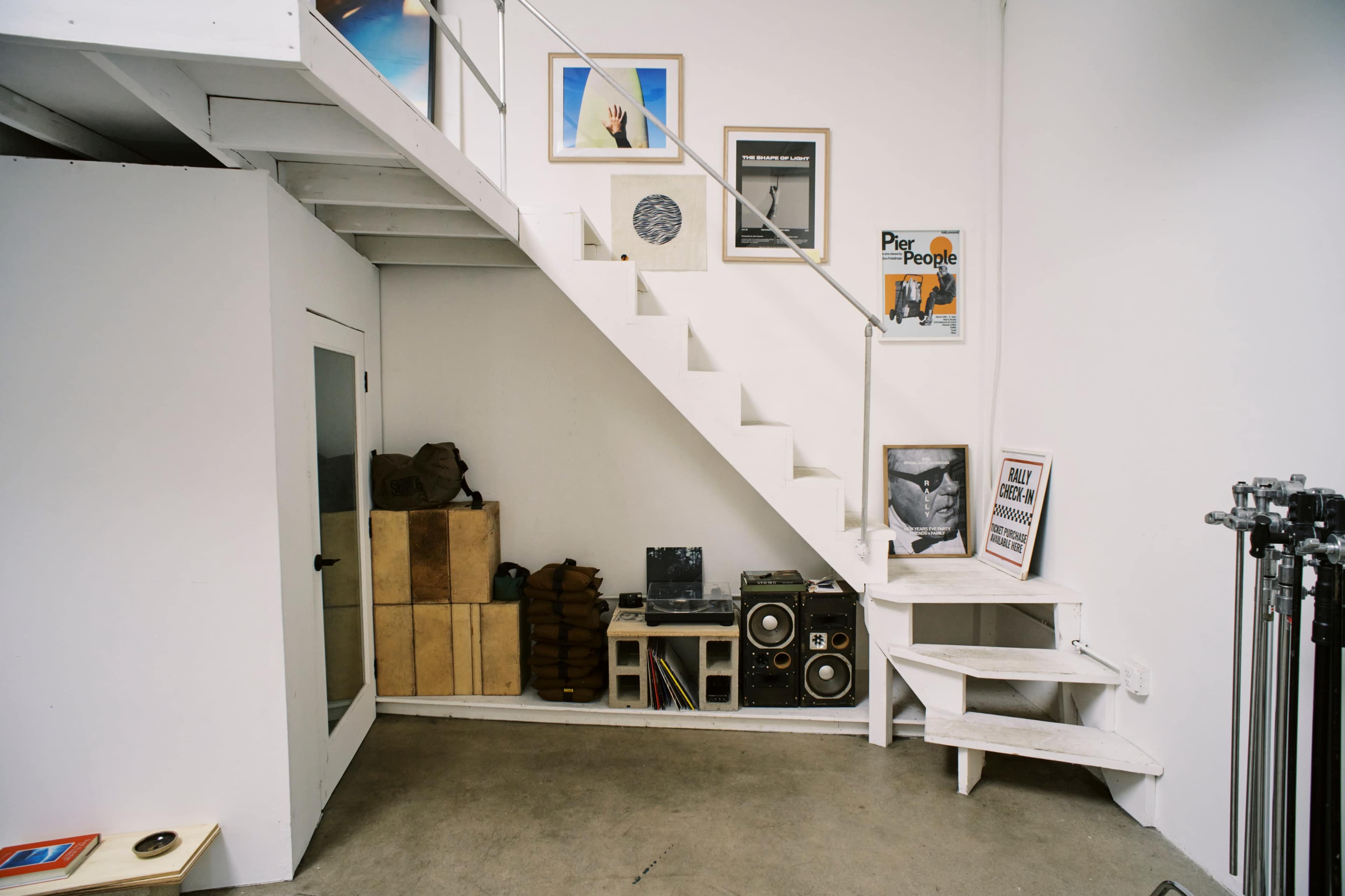 A staircase leads to a small gallery of framed artwork on a white wall adjacent to a storage area with wooden boxes and vintage audio equipment below.