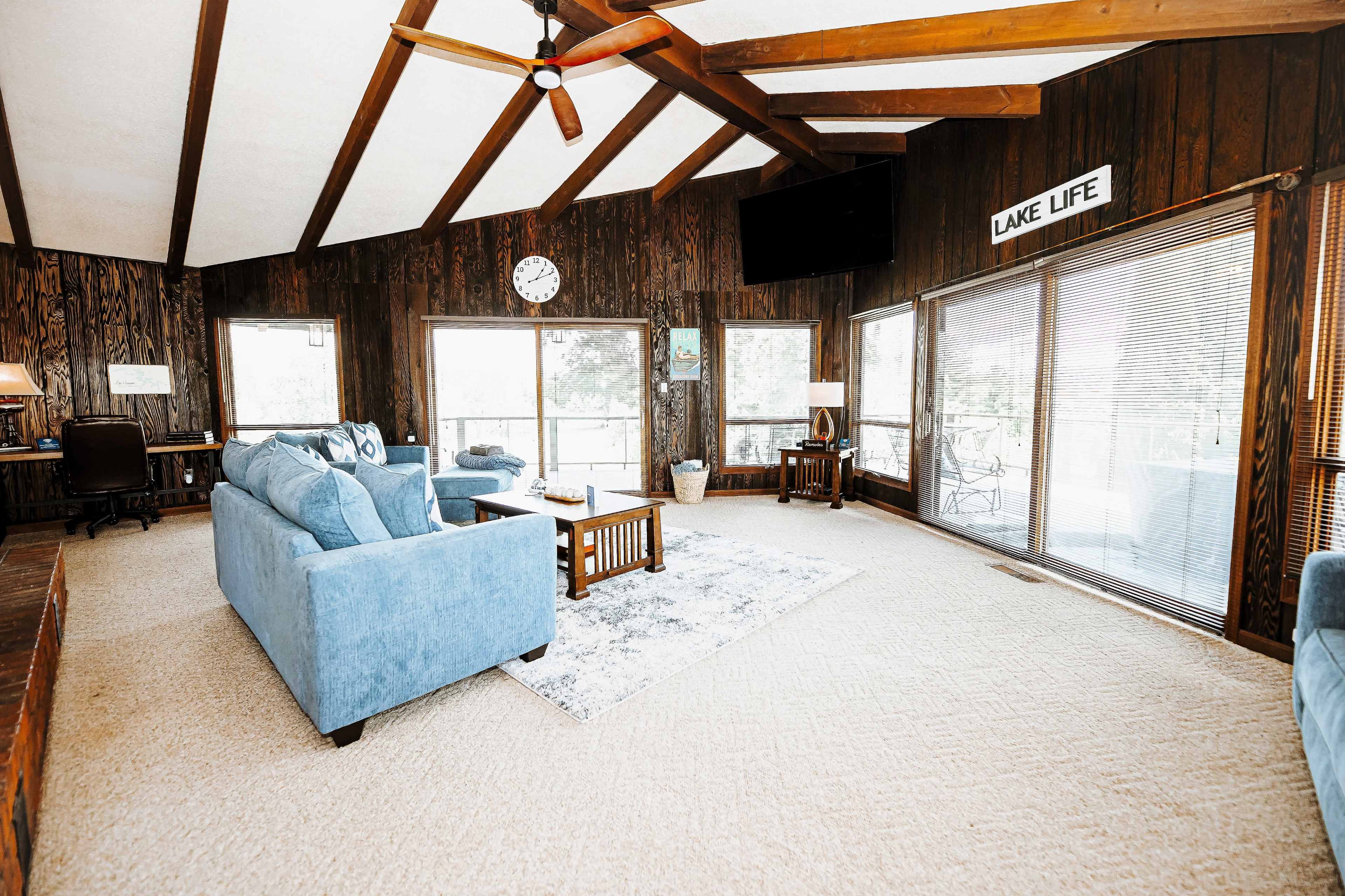 The living room features wooden walls, a high beam ceiling, blue furniture, and large windows overlooking a lake.