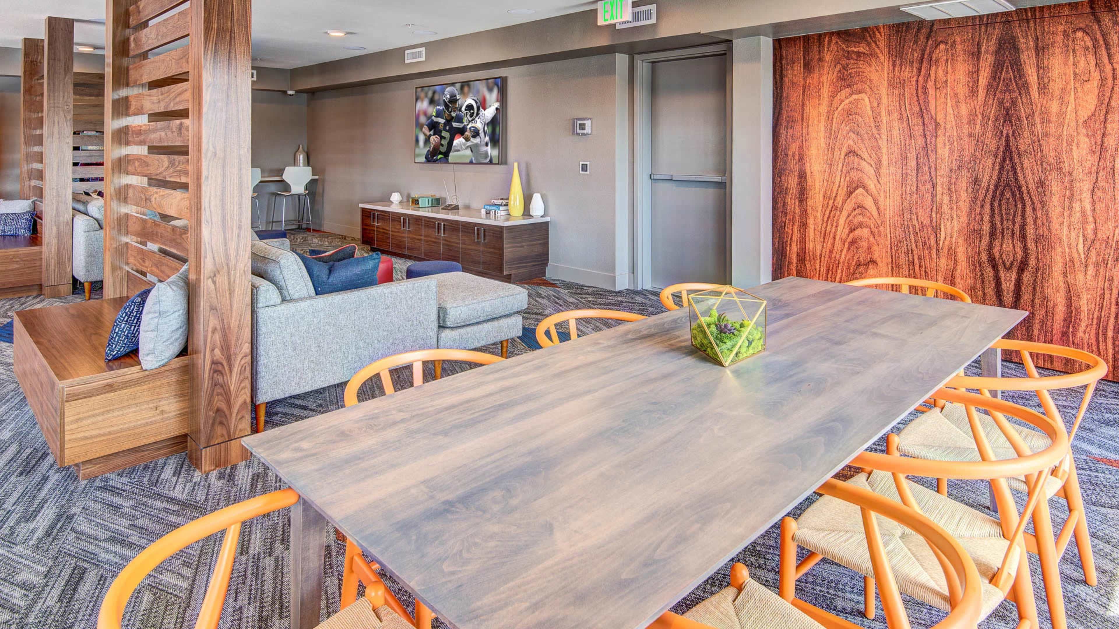 A modern living space features a dining table surrounded by orange chairs, with a couch and a television mounted on the wall in the background.