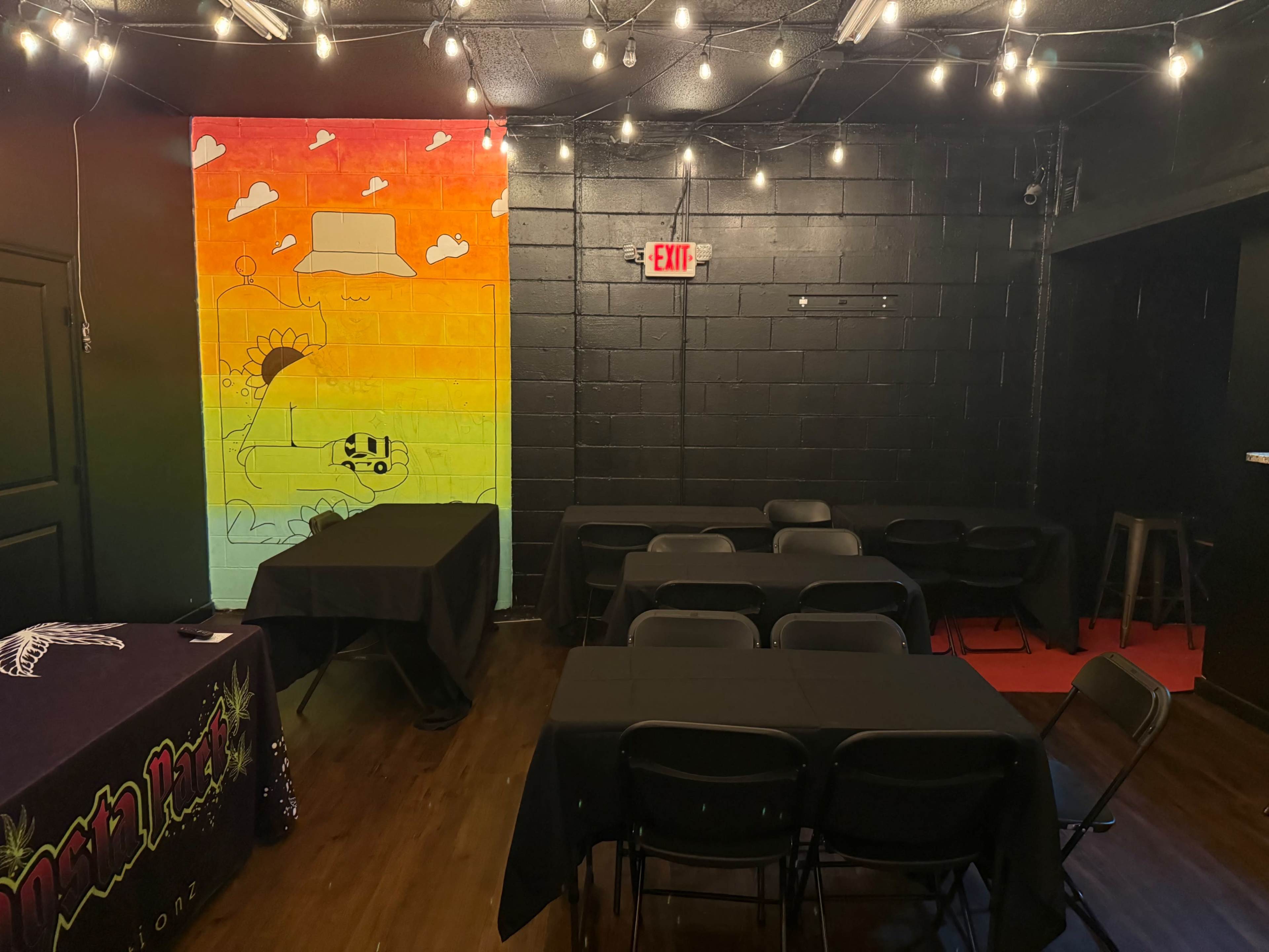 The scene shows a dimly lit room with black walls, featuring tables covered in black cloth and a colorful mural on one wall, illuminated by string lights.