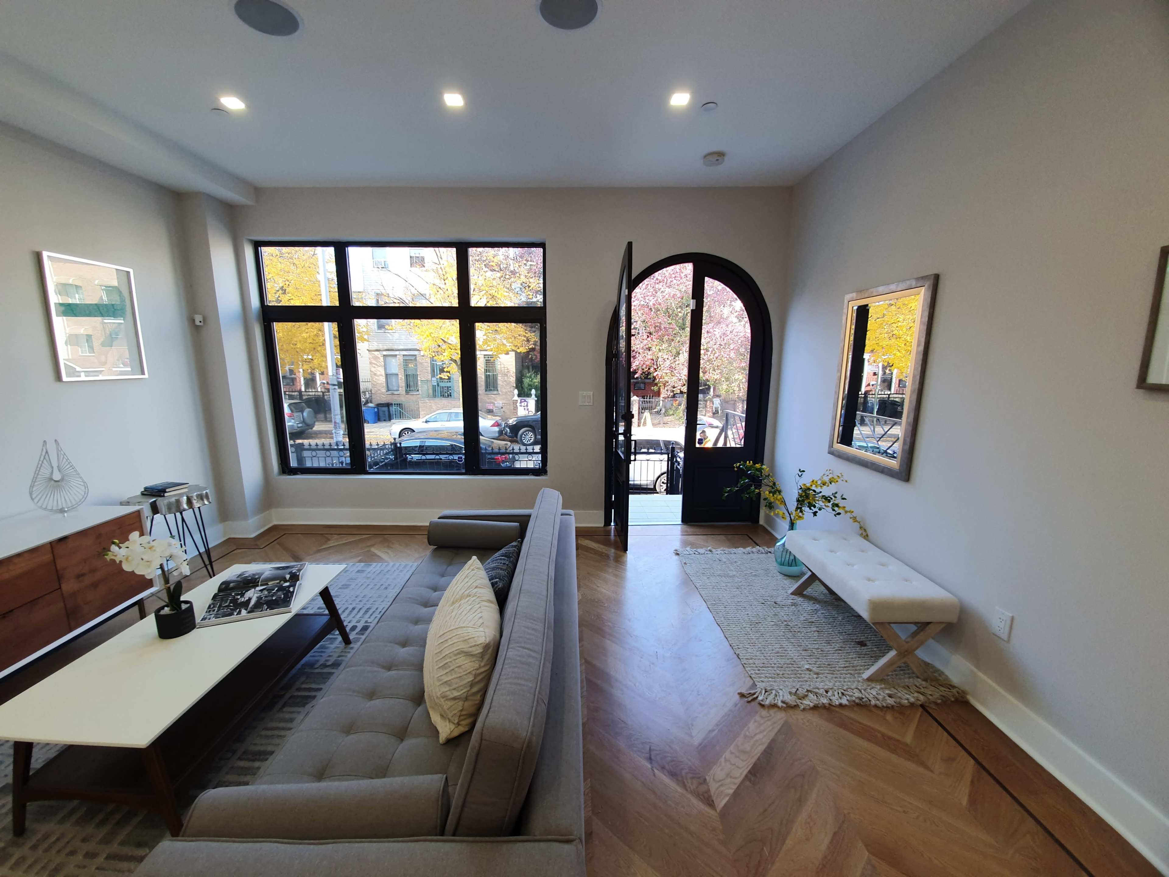 A modern living room features a gray sofa, a coffee table, and large windows that open to a view of a tree-lined street.