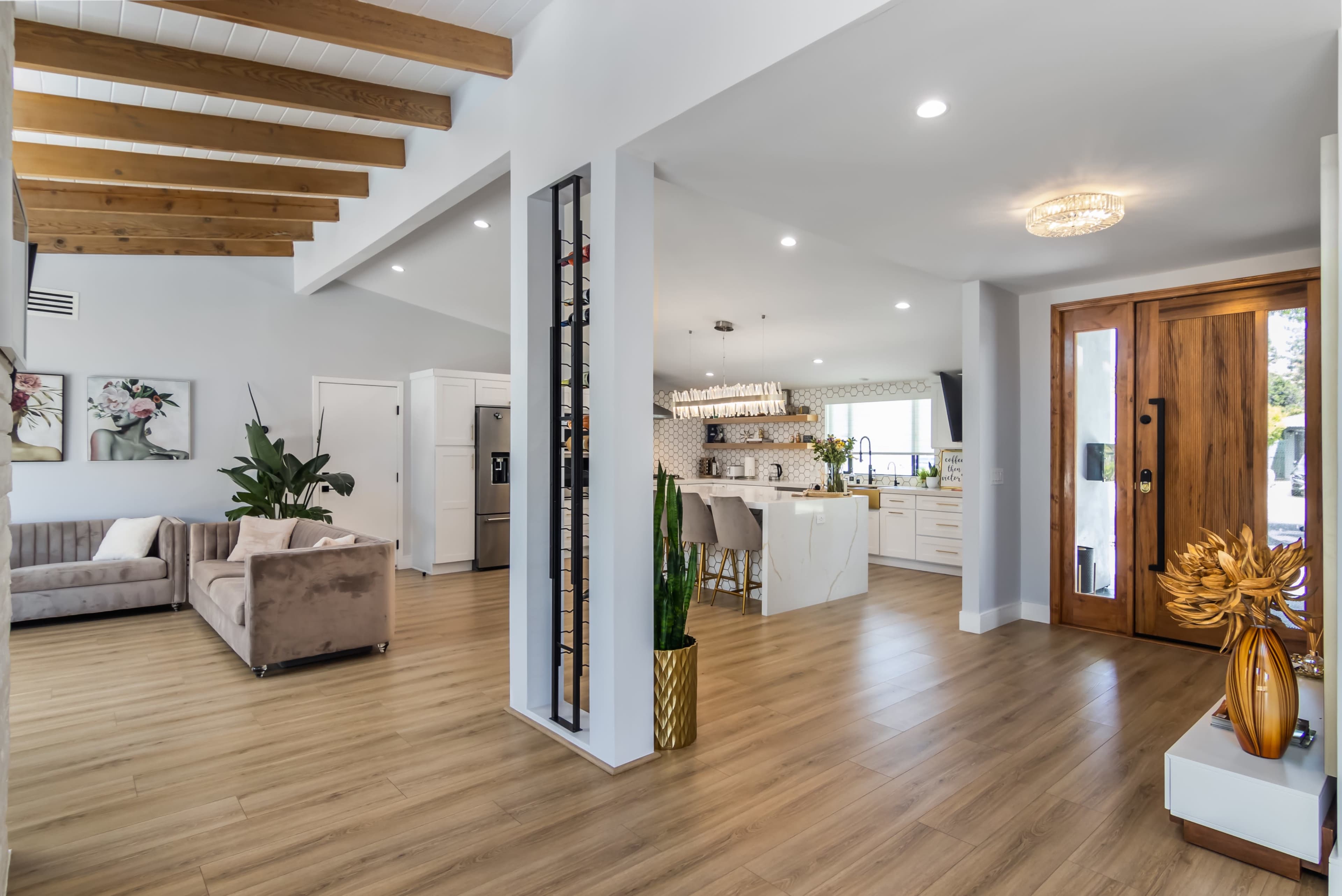 A modern interior with a living area featuring a gray sofa and wooden beams, leading to a kitchen with white cabinetry and a wooden entrance door.