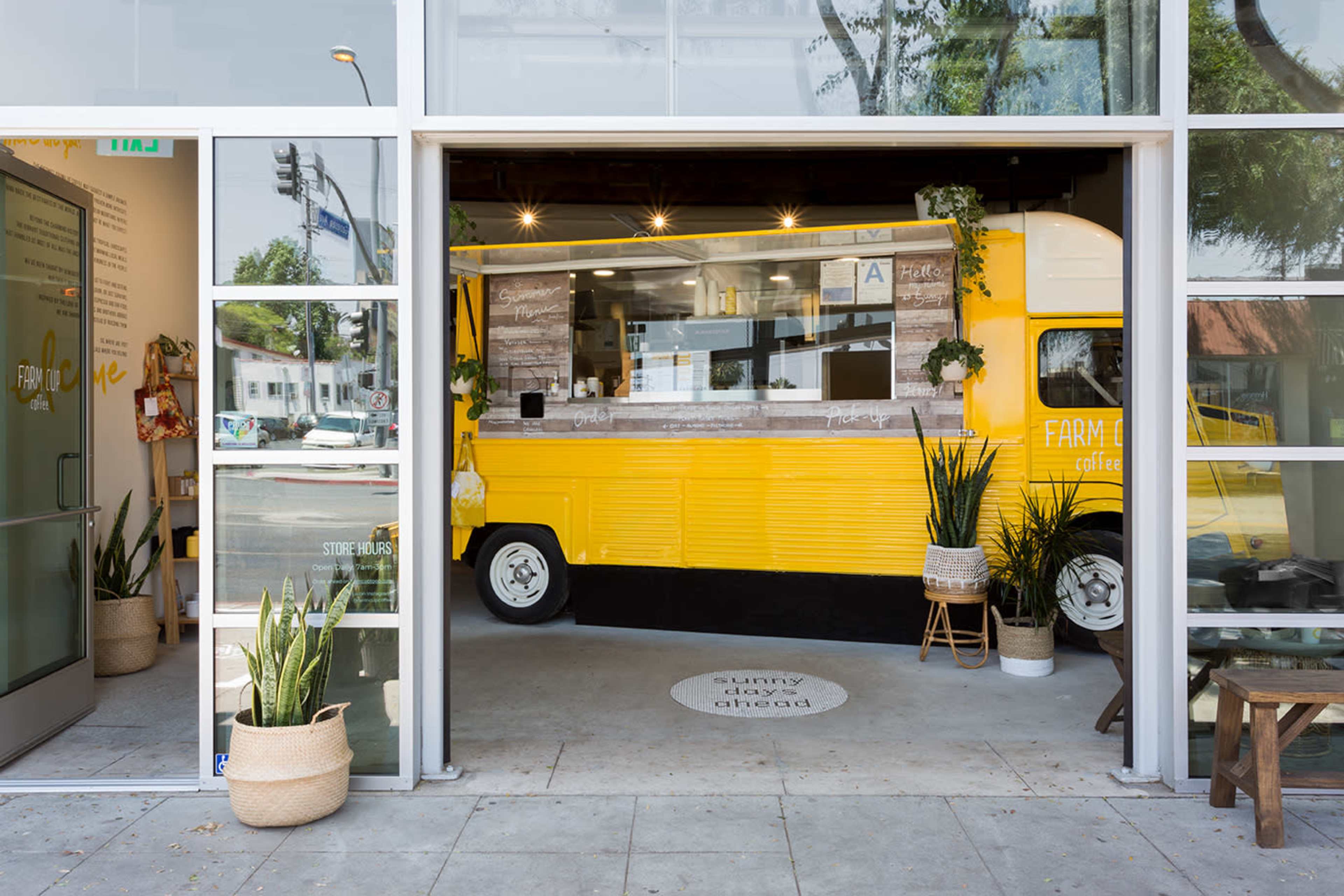 A vibrant yellow food truck is stationed inside a modern café, with potted plants surrounding it and a clear entrance showcasing the menu.